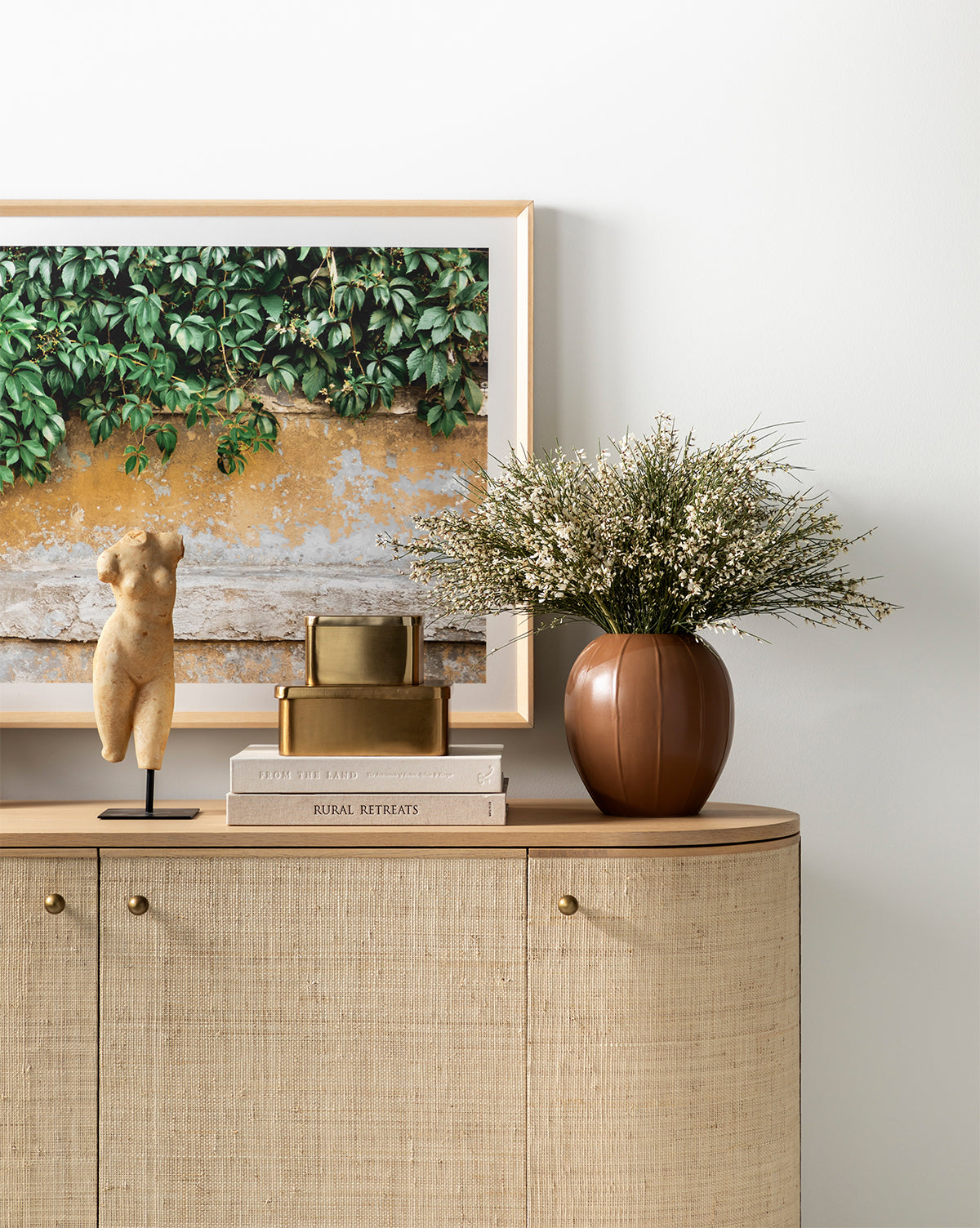 A beige sideboard with gold knobs holds a framed plant photo, a small abstract sculpture, stacked gold boxes, two books, and the brown Idris Vase filled with delicate white flowers against a white wall.