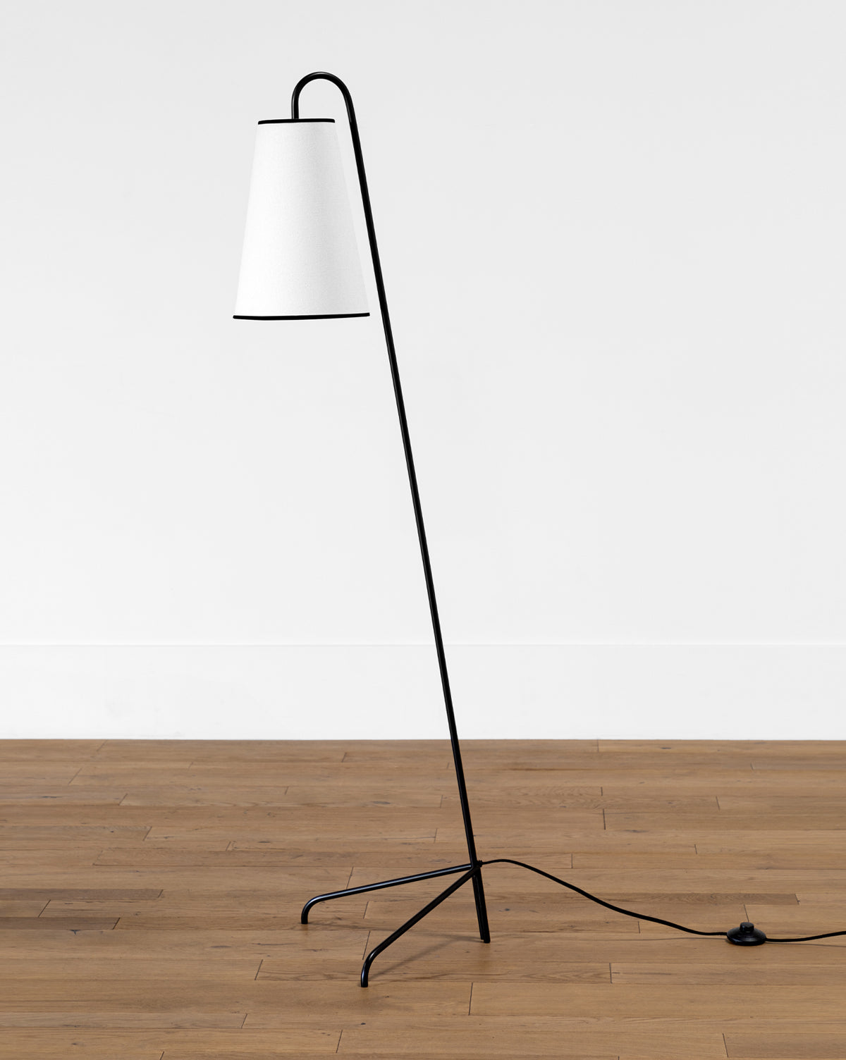 The Makerspalm Jonas Floor Lamp has a slender black metal frame, angled stem, white cone-shaped linen shade, and three-pronged legs, with a visible black power cord for a modern look. McGee & Co.
