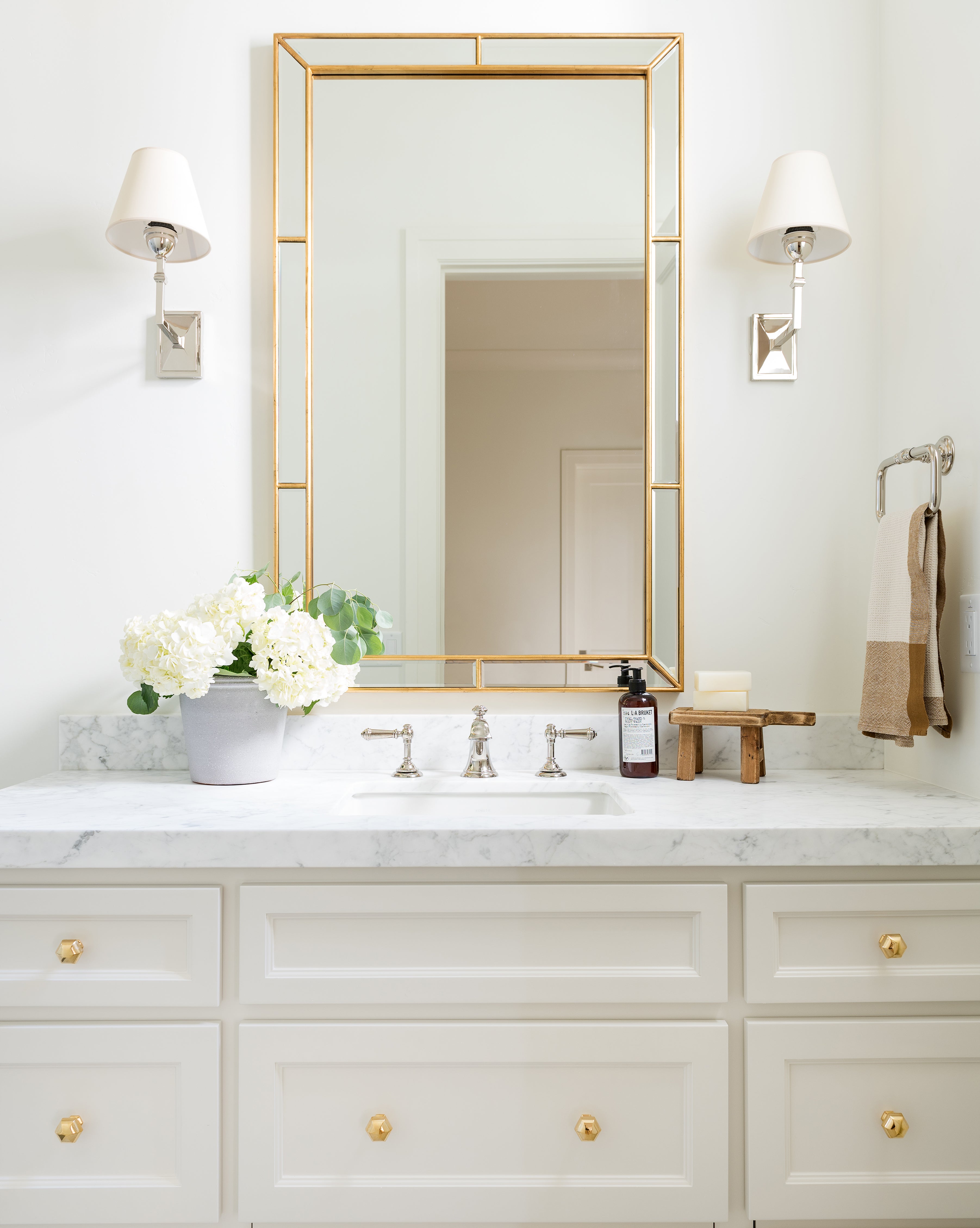 A bright, elegant bathroom features a marble countertop with a potted white hydrangea. Above the sink, a Celine Mirror with beveled glass and a burnished gold leaf frame is flanked by two sconces with white shades; a towel hangs nearby, by McGee & Co.