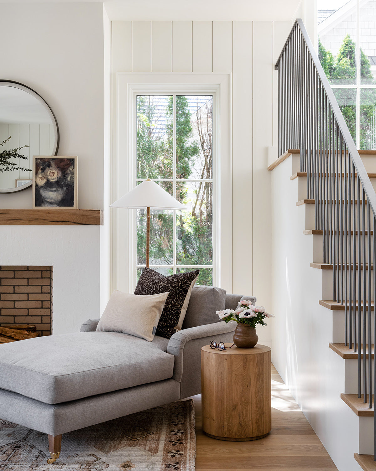 A cozy living space features the Community Lucille English Roll Arm Chaise Lounge with a down-wrapped cushion, a wooden side table with flowers and glasses, a brick fireplace topped by art and a round mirror, plus black-railed stairs beside a large window—McGee & Co.