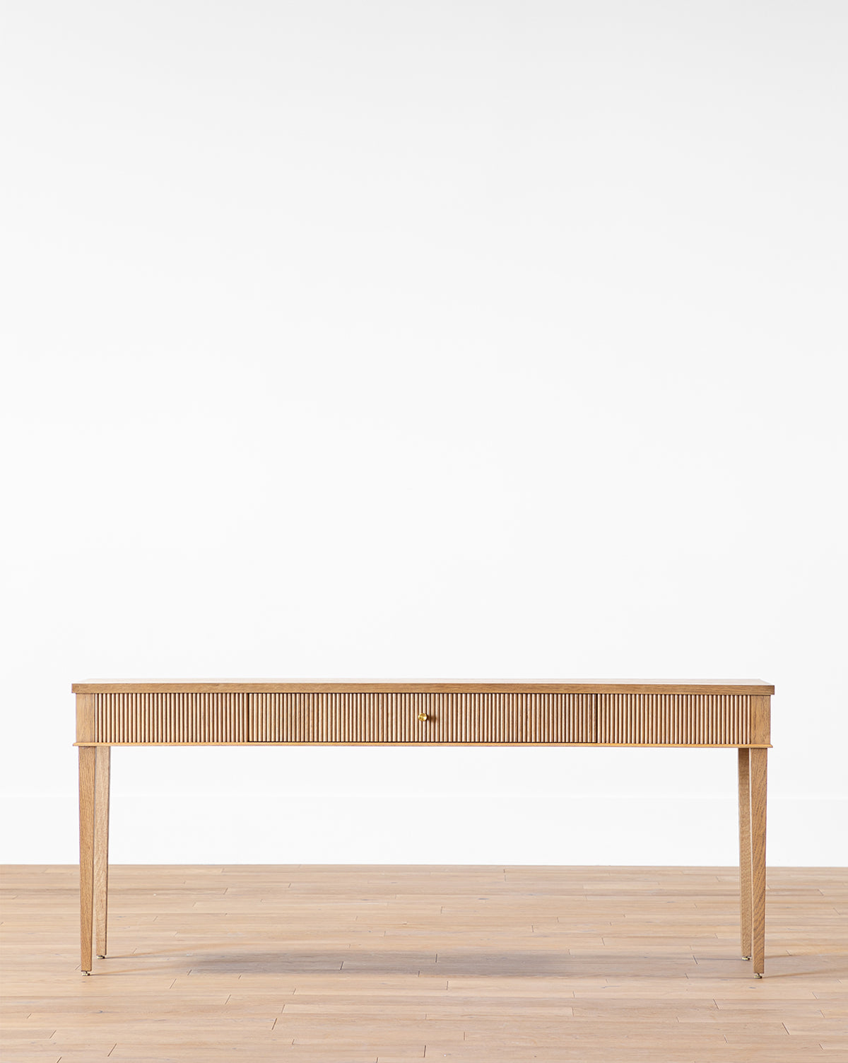 The Pieter Console, a minimalist wooden piece with a concealed drawer, four tapered legs, and subtle reeded detail, stands on a light wood floor against a plain white wall by McGee & Co.