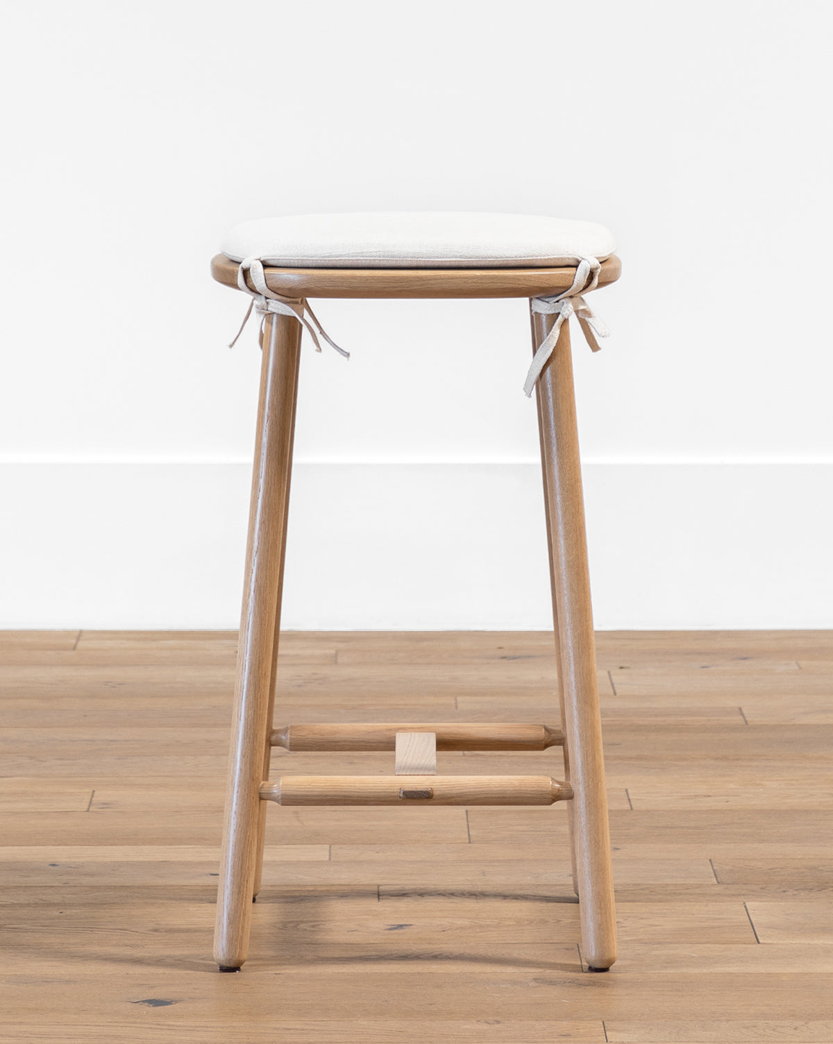The Ralph Oak Counter Stool features a wooden oak frame and a light linen cushion tied at the corners, placed on a wooden floor against a white wall by McGee & Co.