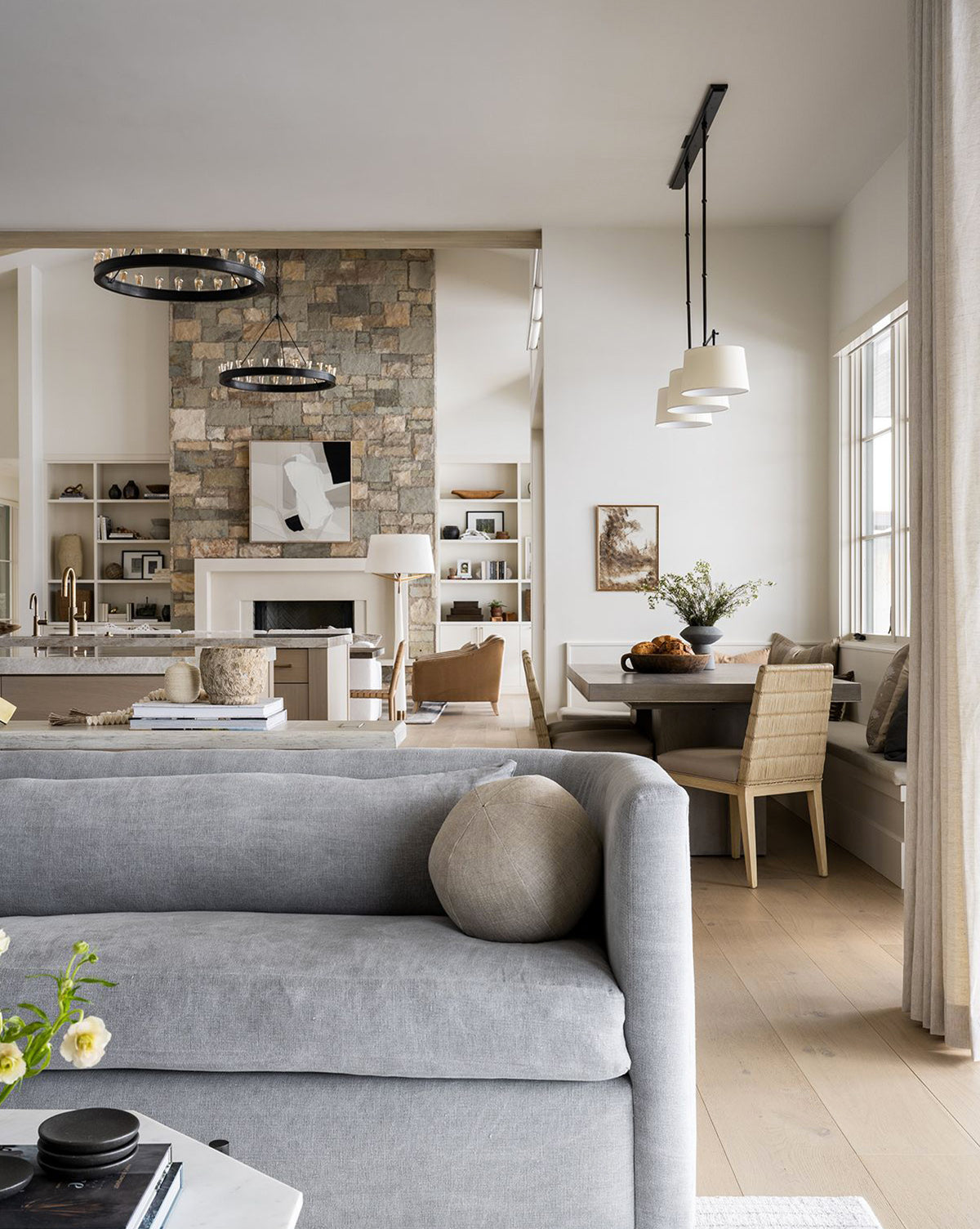 A modern, bright living area showcases a Reese Sofa (84-96) with down-wrapped cushions, a stone fireplace, handcrafted alder wood shelves in the kitchen, a round dining table with chairs, and large windows bringing in natural light by McGee & Co.