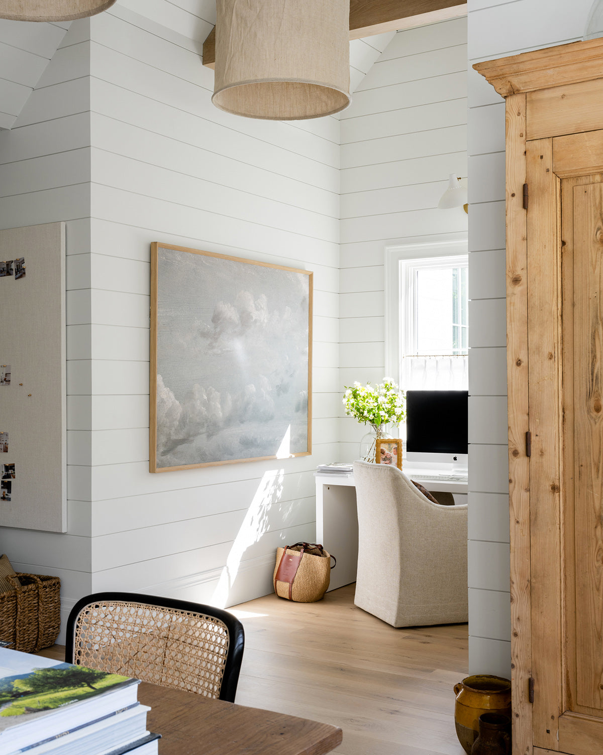 A cozy home office features white shiplap walls, a large Gray Clouds framed wall art, a beige armchair at a white computer desk, and sunlight streaming through the window with flowers for relaxed decor.