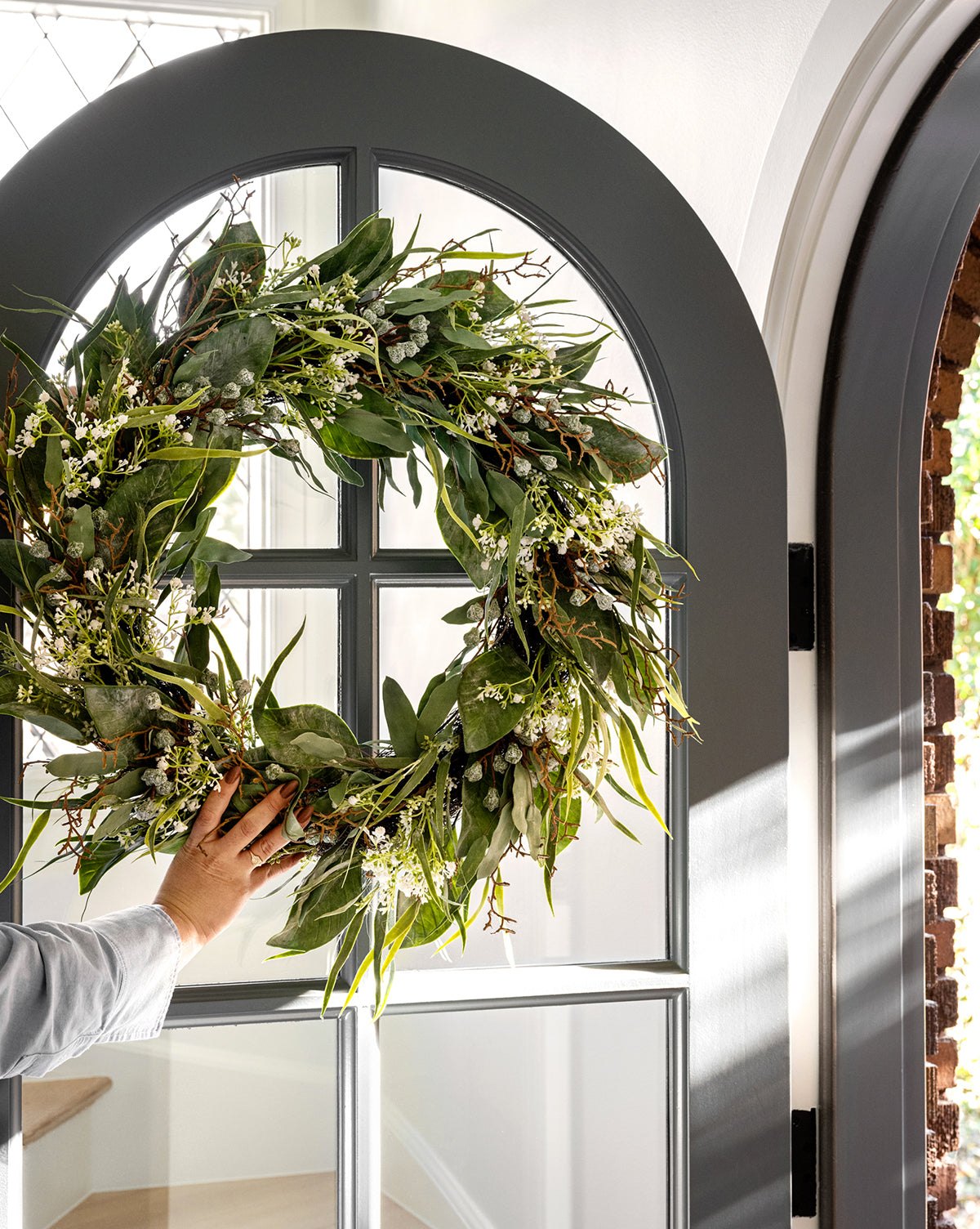 A hand hangs the Faux Wild Grass & Eucalyptus Wreath, featuring small white flowers, on a glass-paneled arched gray door in a sunlit entryway.