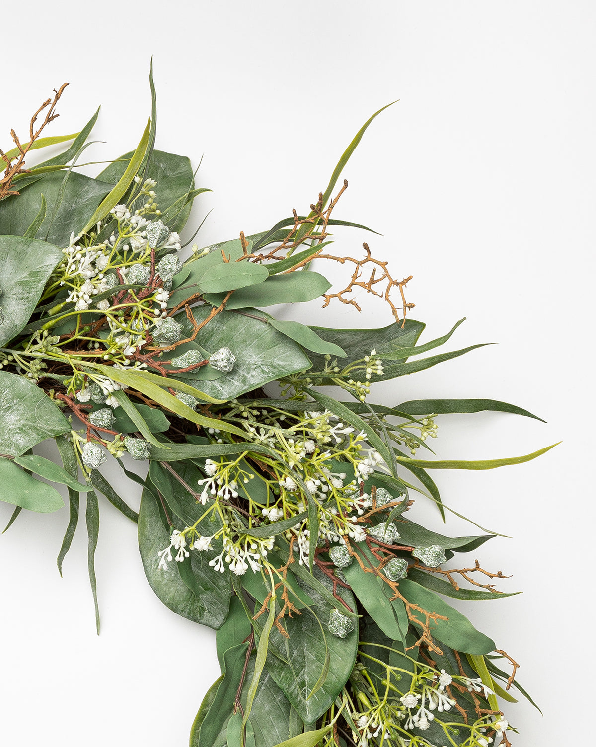 A close-up of the Faux Wild Grass & Eucalyptus Wreath, featuring faux greenery with green leaves, small white flowers, and brown branches, displayed against a plain white background.