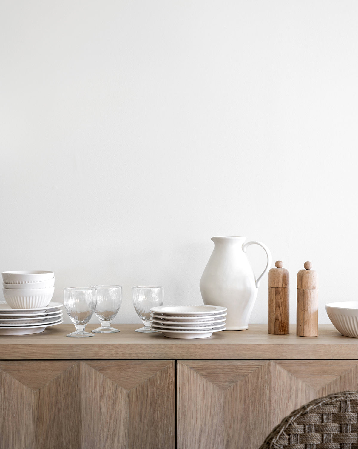 A minimalist wooden sideboard with fluted detailing displays stacked white dishes, clear glasses, a white ceramic pitcher, the Adele Bowl, and wooden salt and pepper grinders against a plain white wall, by McGee & Co.