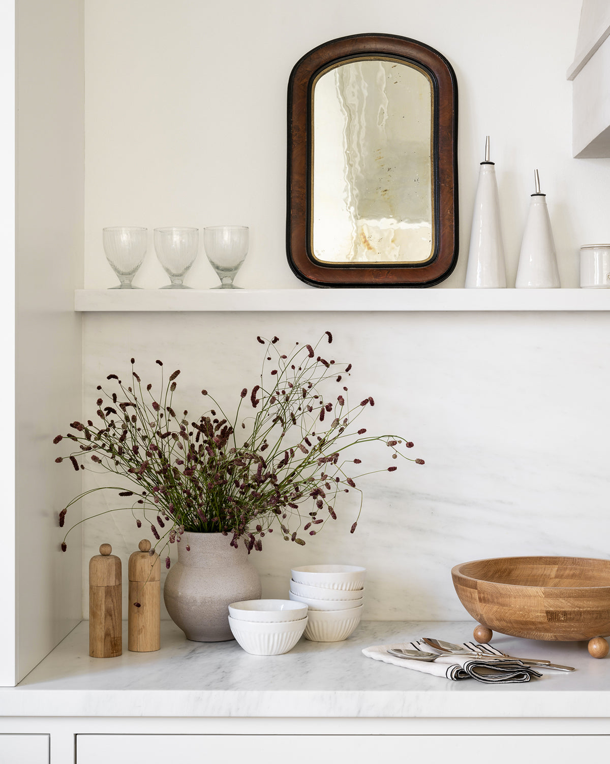 A minimalist kitchen shelf features a rustic mirror, fluted glassware, tall ceramic bottles, wildflowers in a vase, wooden bowls, stacked Adele Bowls in white, salt and pepper mills, and arranged cutlery on a marble countertop by McGee & Co.