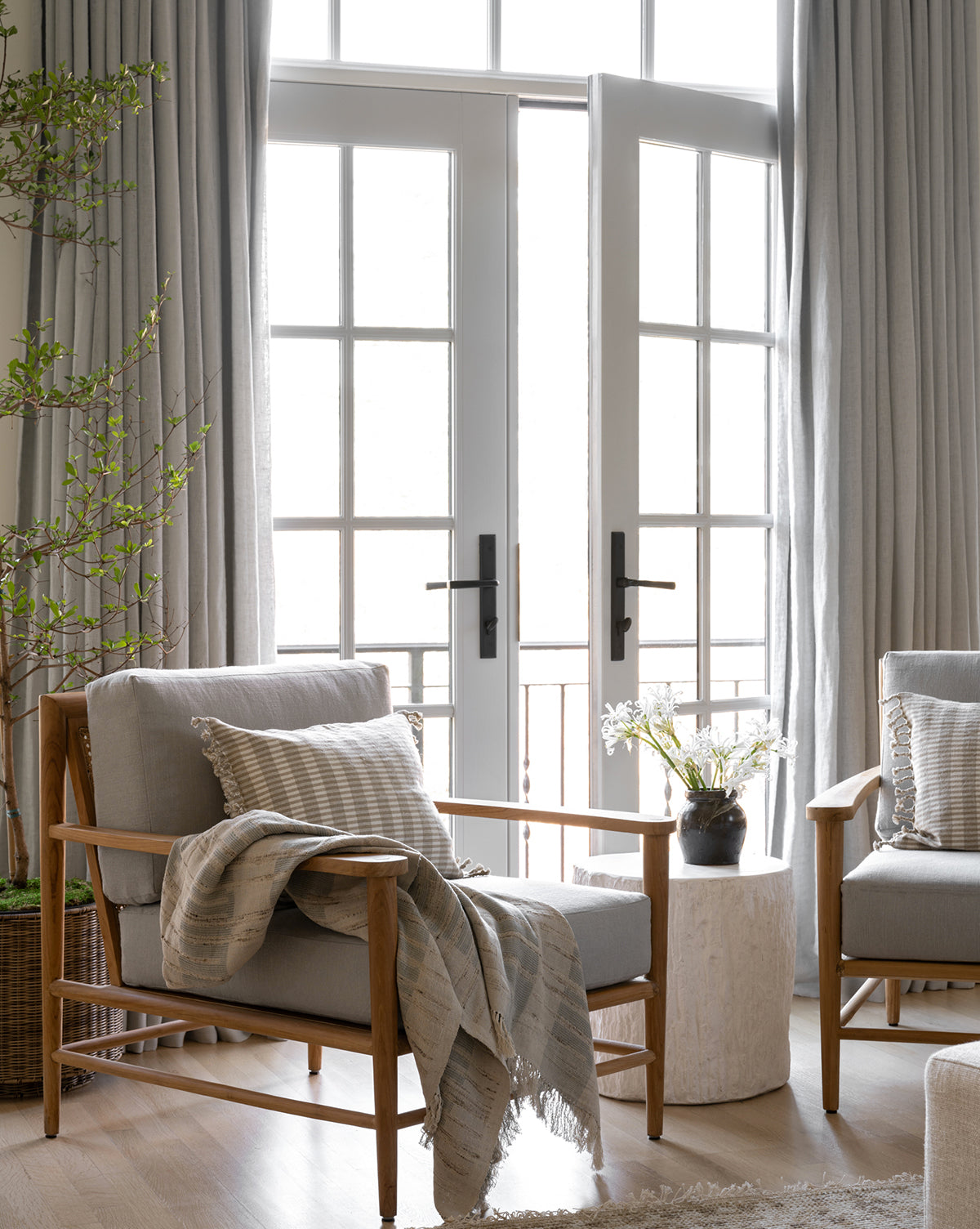 A bright, cozy living room features two wooden armchairs with gray cushions, a textured Theodora Block Stripe Throw, pillows, a round white side table with a vase of flowers, tall curtains, and French doors letting in natural light.