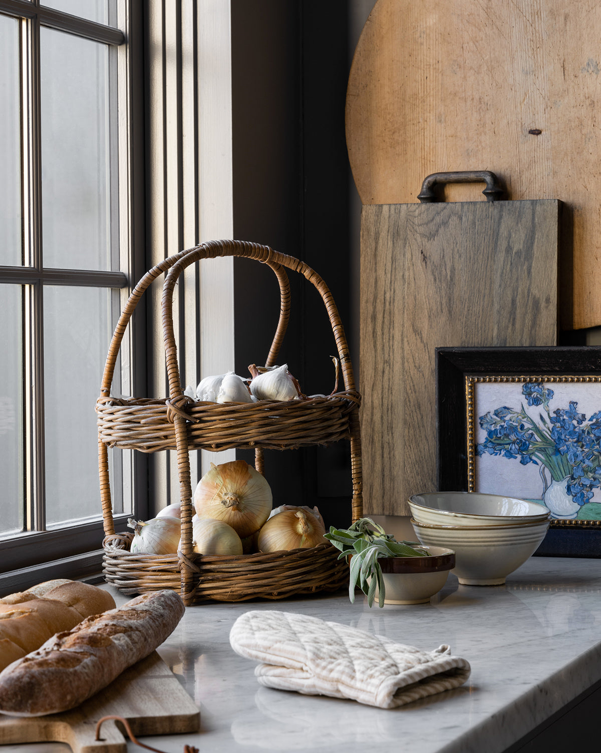 A kitchen countertop displays a two-tiered wicker basket with onions and garlic, a loaf of bread, the Fordham Board (solid oak), ceramic bowls, floral artwork in a frame, and a white oven mitt by the window, by McGee & Co.