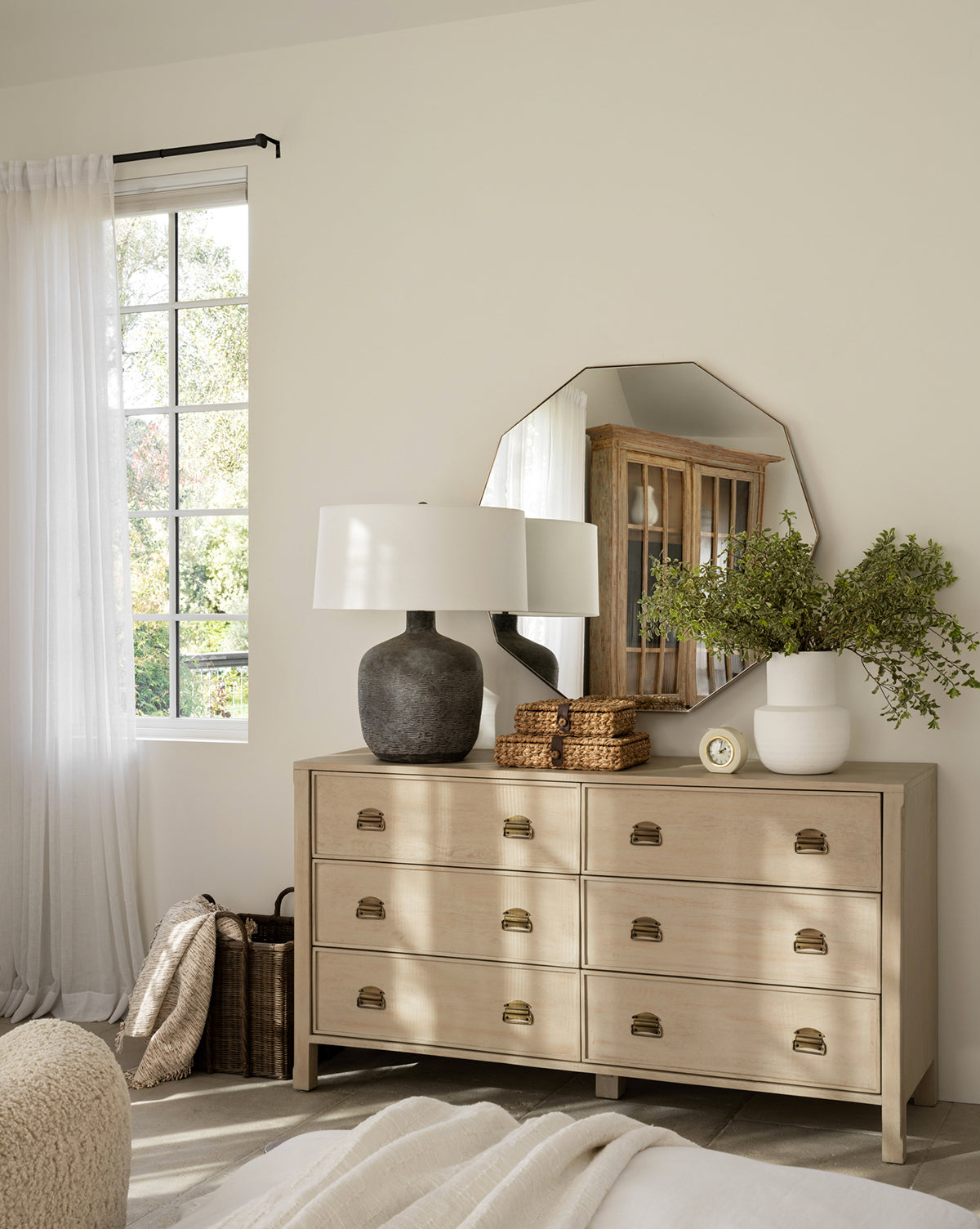 A light wood dresser with brass handles stands by a cream wall, topped with the Hawley Decagon Mirror, two vases (one filled with greenery), baskets, a lamp, and a clock, by McGee & Co. Sunlight filters in through sheer curtains.