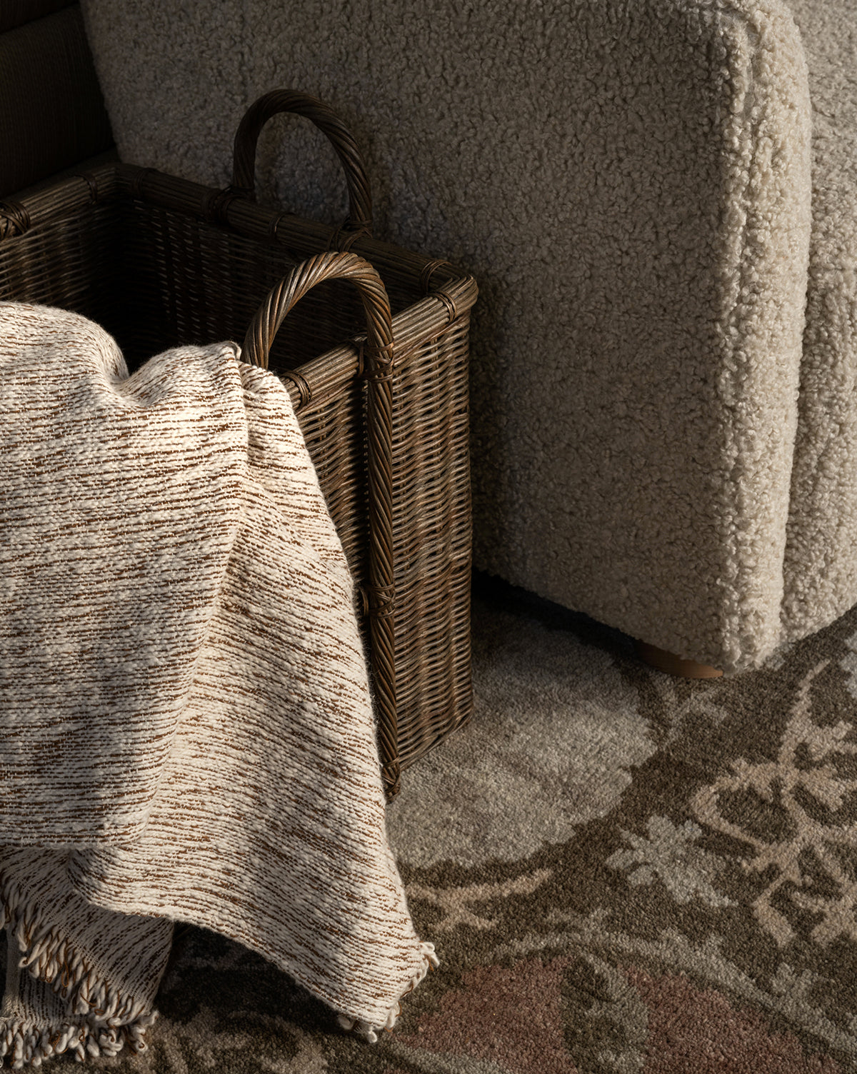 A textured blanket drapes over the side of the Jacinta Woven Basket, adding organic texture beside a beige boucle upholstered chair on a patterned rug.