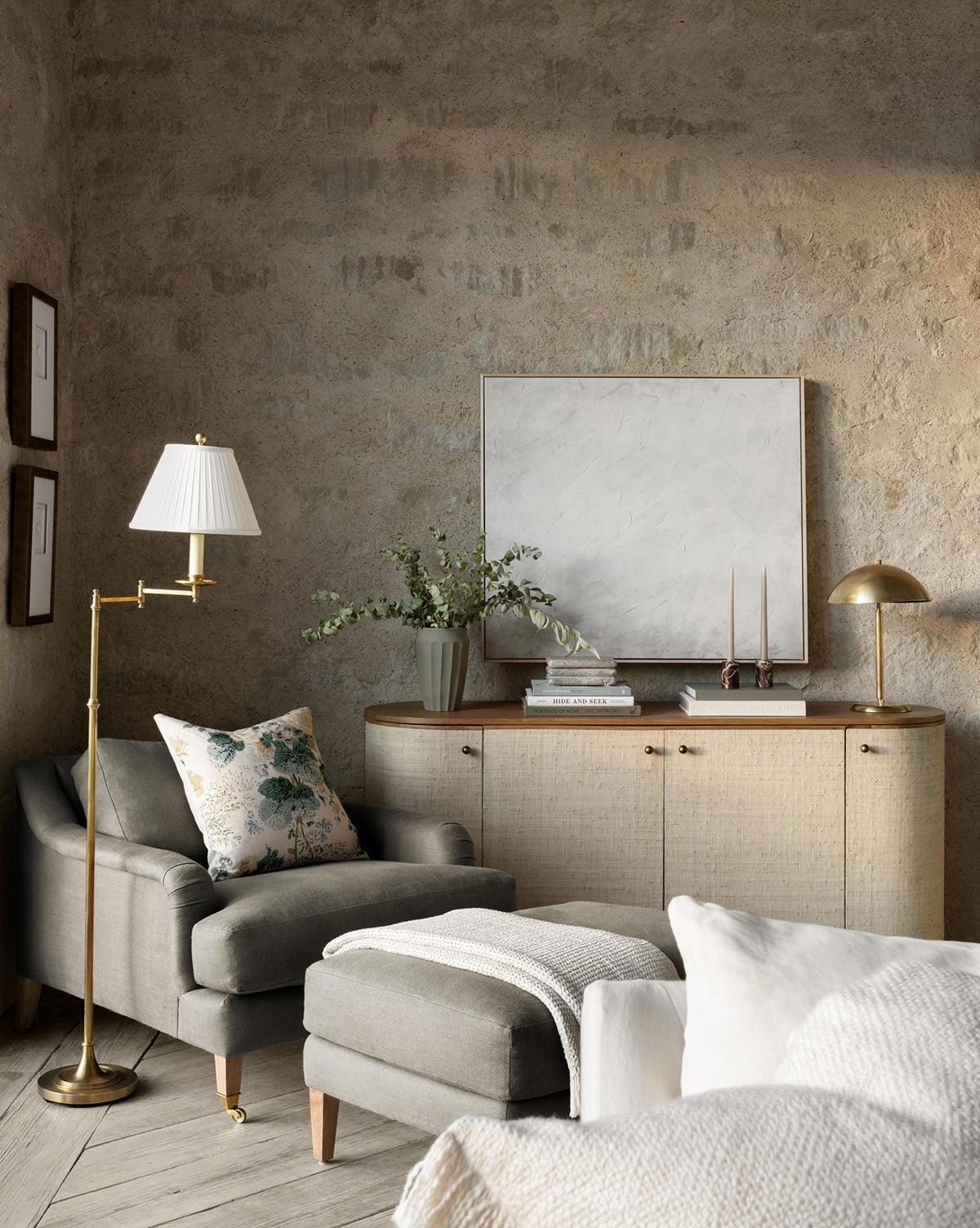 A cozy living room corner showcases the Community Lucille English Roll Arm Lounge Chair with a down-wrapped cushion, matching ottoman, floral pillow, floor lamp, light wood sideboard with decor, framed artwork, and a neutral textured wall—all featuring pieces from McGee & Co.