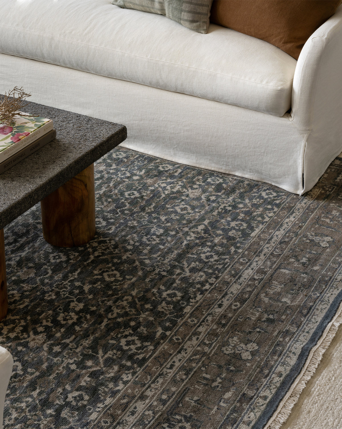 A close-up of a living room featuring the Marta Hand-Knotted Wool Rug, a granite coffee table with a book, and a white sofa adorned with beige and brown throw pillows, by McGee & Co.