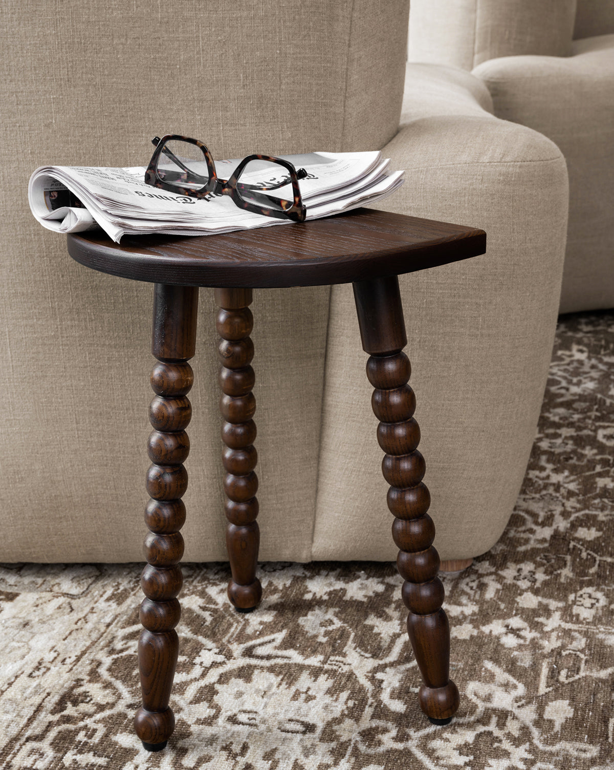 The Alice Wood Stool with three beaded ash wood legs holds a folded newspaper and eyeglasses beside a beige upholstered sofa on a patterned rug, by McGee & Co.