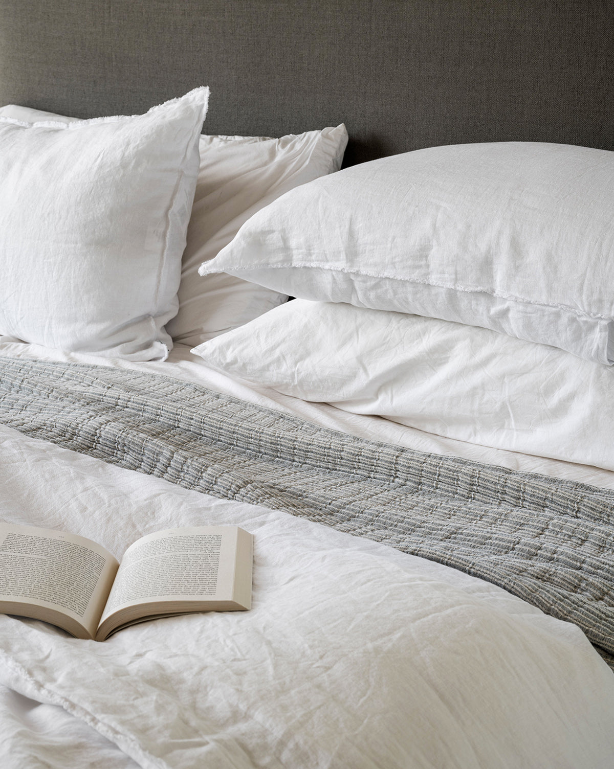 A neatly made bed with white pillows and bedding, accented by the Hanson Striped Cotton Coverlet at the foot and an open book on the sheets, creates a cozy showcase of modern bedding styles.
