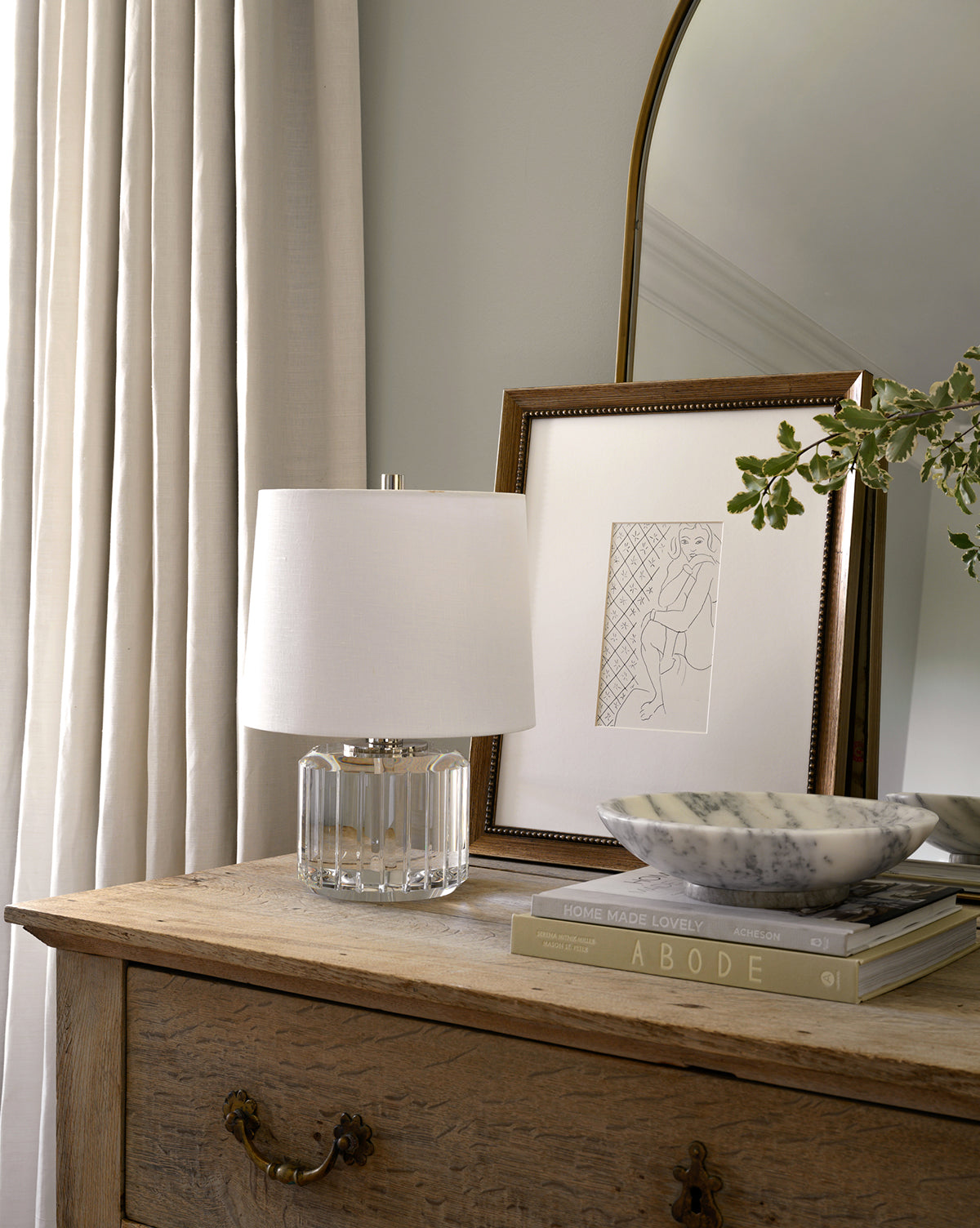 A wooden dresser with brass handles displays a crystal lamp with a white shade, a framed minimalist line drawing, the Arabesco Marble Bowl, and two stacked books next to cream curtains and a large mirror.