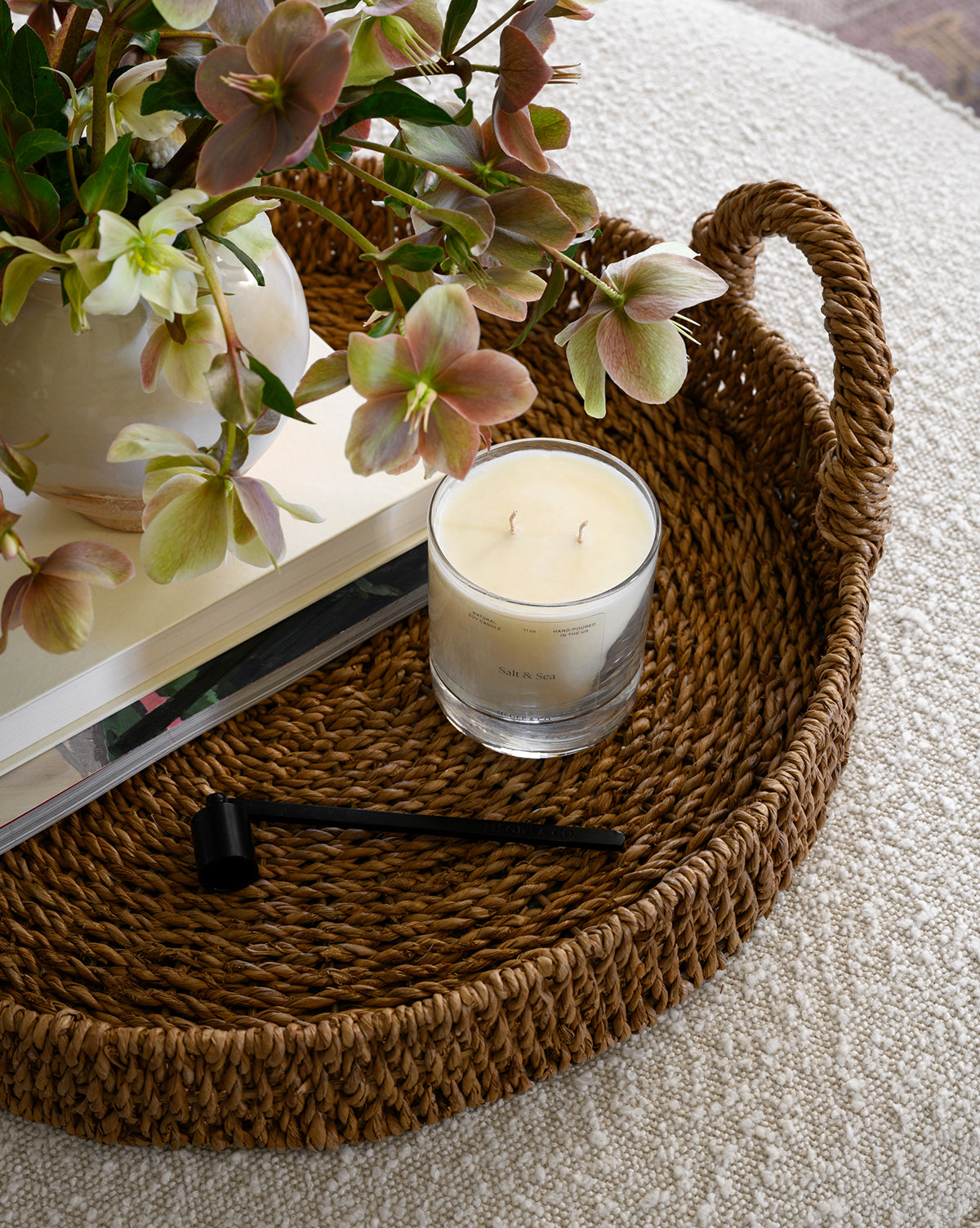 A woven wicker tray displays a glass candle, the Candle Snuffer, a stack of books, and a vase with green and pink flowers, all arranged on a textured light surface.