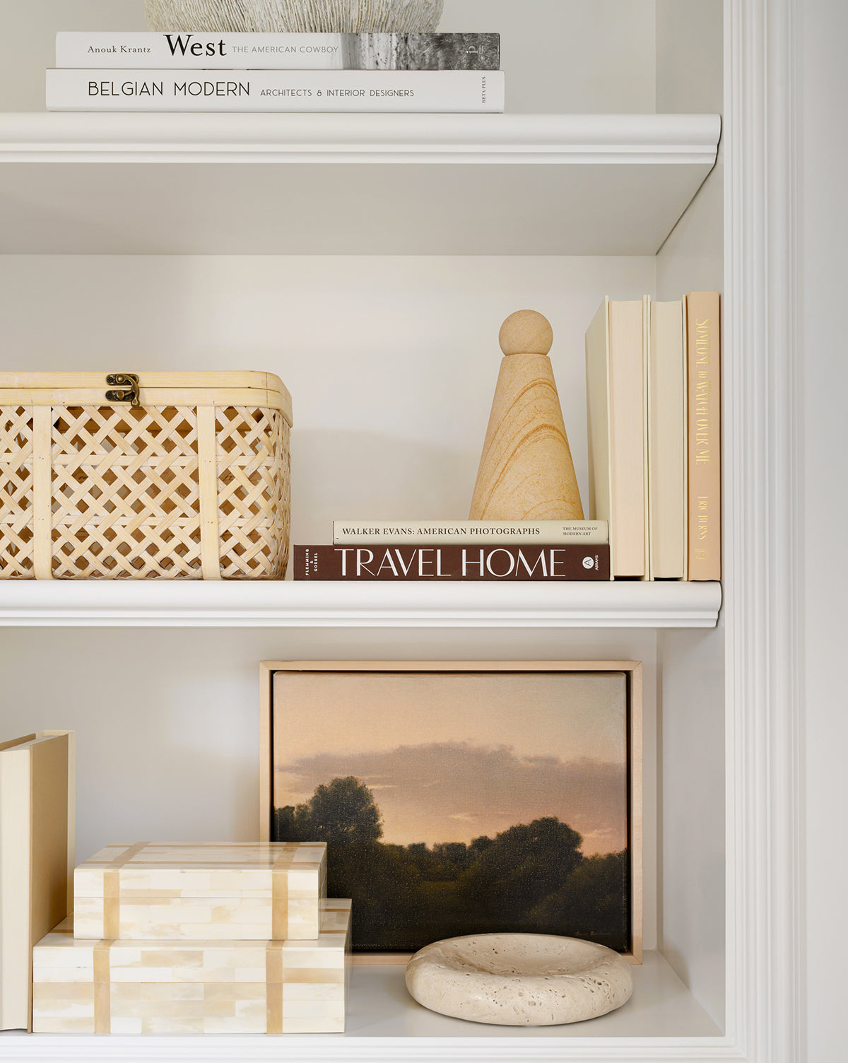 A neatly styled white bookshelf displays stacked books, a woven basket, decorative boxes, a round stone, a wooden chess piece, and framed wall art titled The Grove by Bruce Brainard, featuring a meditative landscape in warm tones.