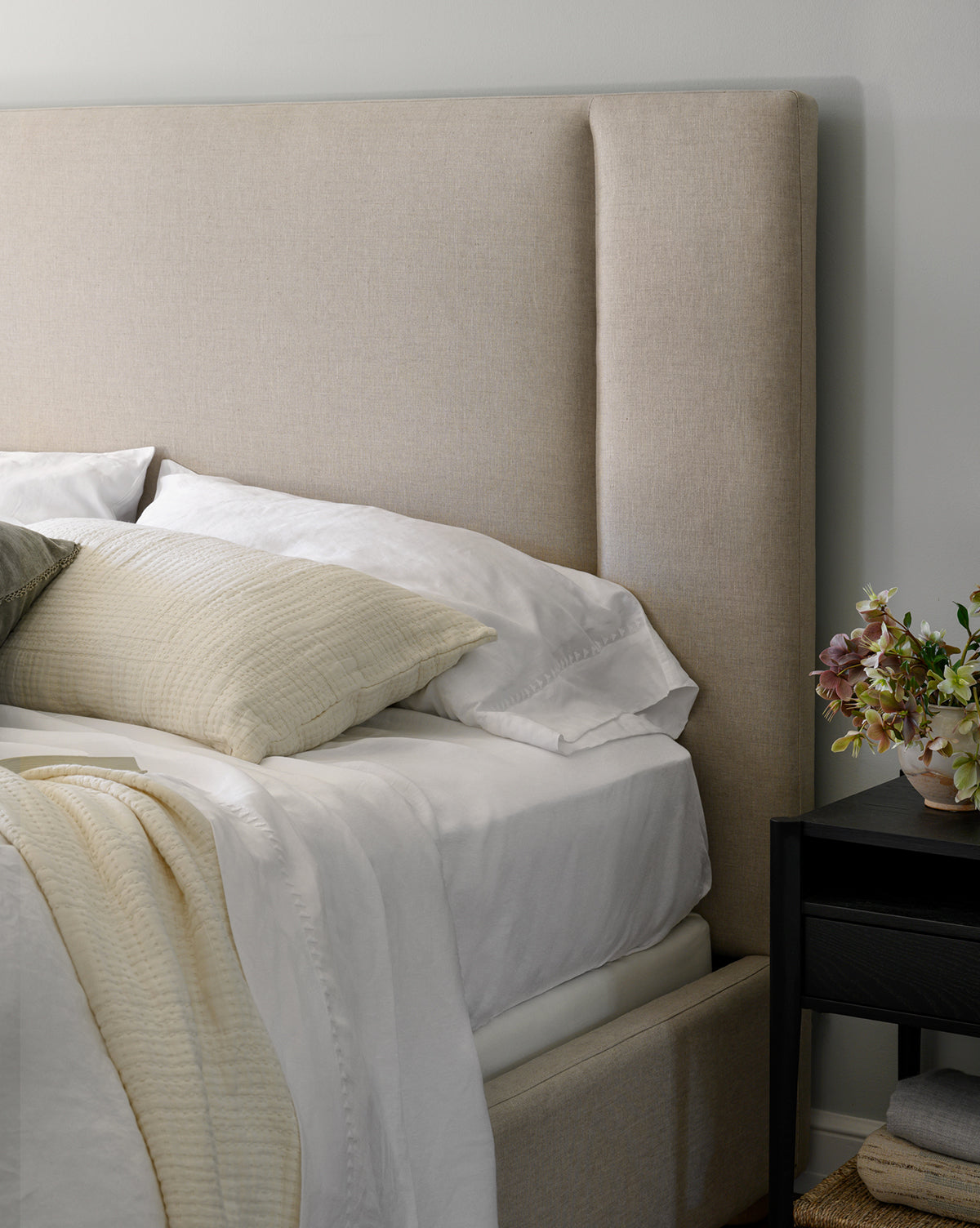 The Mina Bed features a beige upholstered frame, premium fabrics, white sheets, and cream pillows. It sits next to a black nightstand topped with a small floral arrangement in a vase, by McGee & Co.