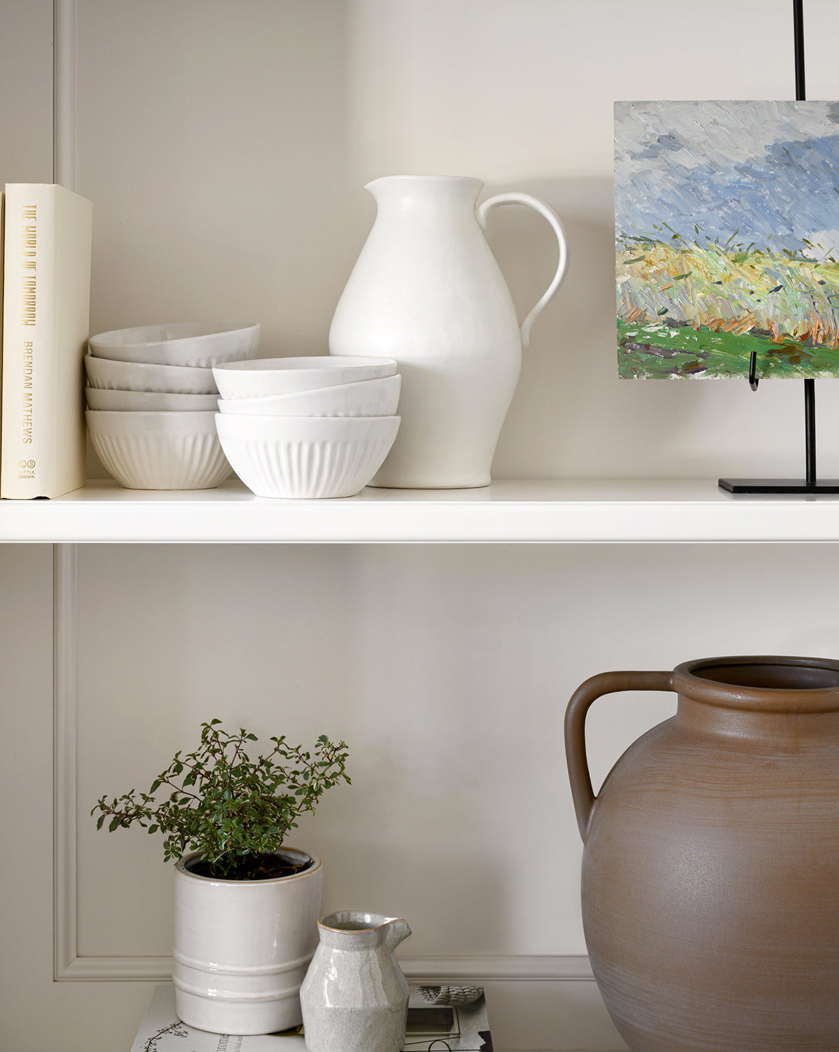 Two white shelves showcase stacked Adele Bowls with fluted detailing, a white pitcher, a small canvas painting on a stand, a potted plant, a grayish jug, and a large brown pot. A book stands upright at the left end, by McGee & Co.