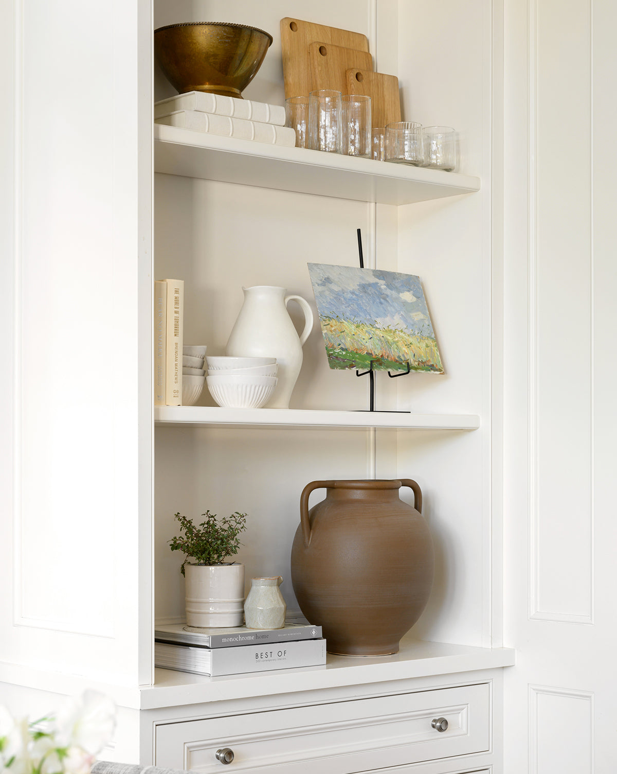 White built-in shelves display stacked books, glassware, white ceramic dishes, a jug, a Rounded Dual Handled Vase in soft brown tones with a tapered base, a small potted plant, and an art print on a stand in a clean, minimalist space.