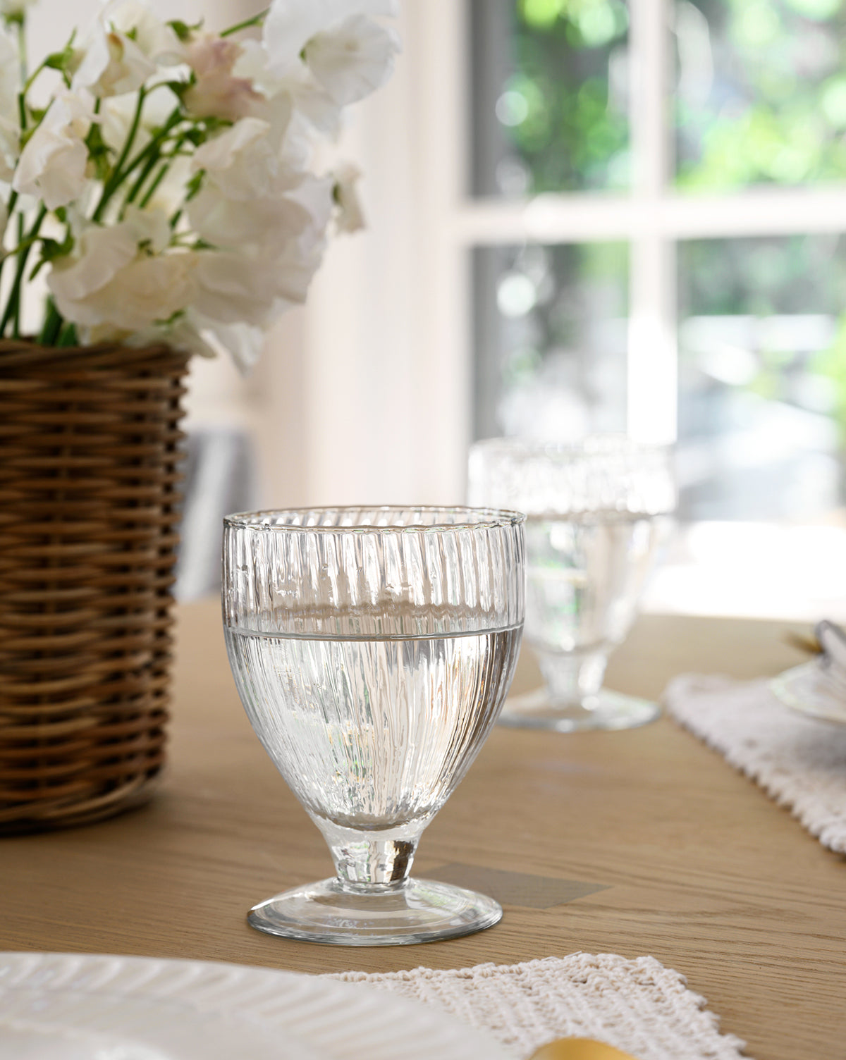 A Ribbed Wine Glasses (Set of 4) sits on a wooden table, filled with water, beside white flowers in a wicker basket as sunlight streams through the window in the background, by McGee & Co.