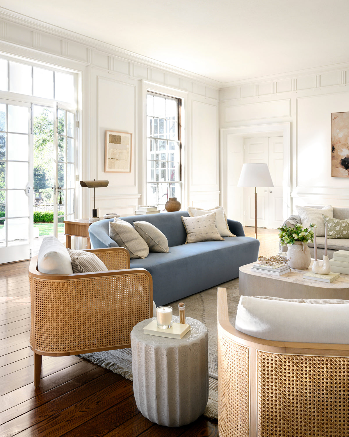 Bright living room with large windows, a light blue sofa, wooden floors, neutral décor, and natural details. Sunlight highlights cozy cushions, plants, art, and the inviting Elowyn Lounge Chair with caned accents for a serene atmosphere by McGee & Co.