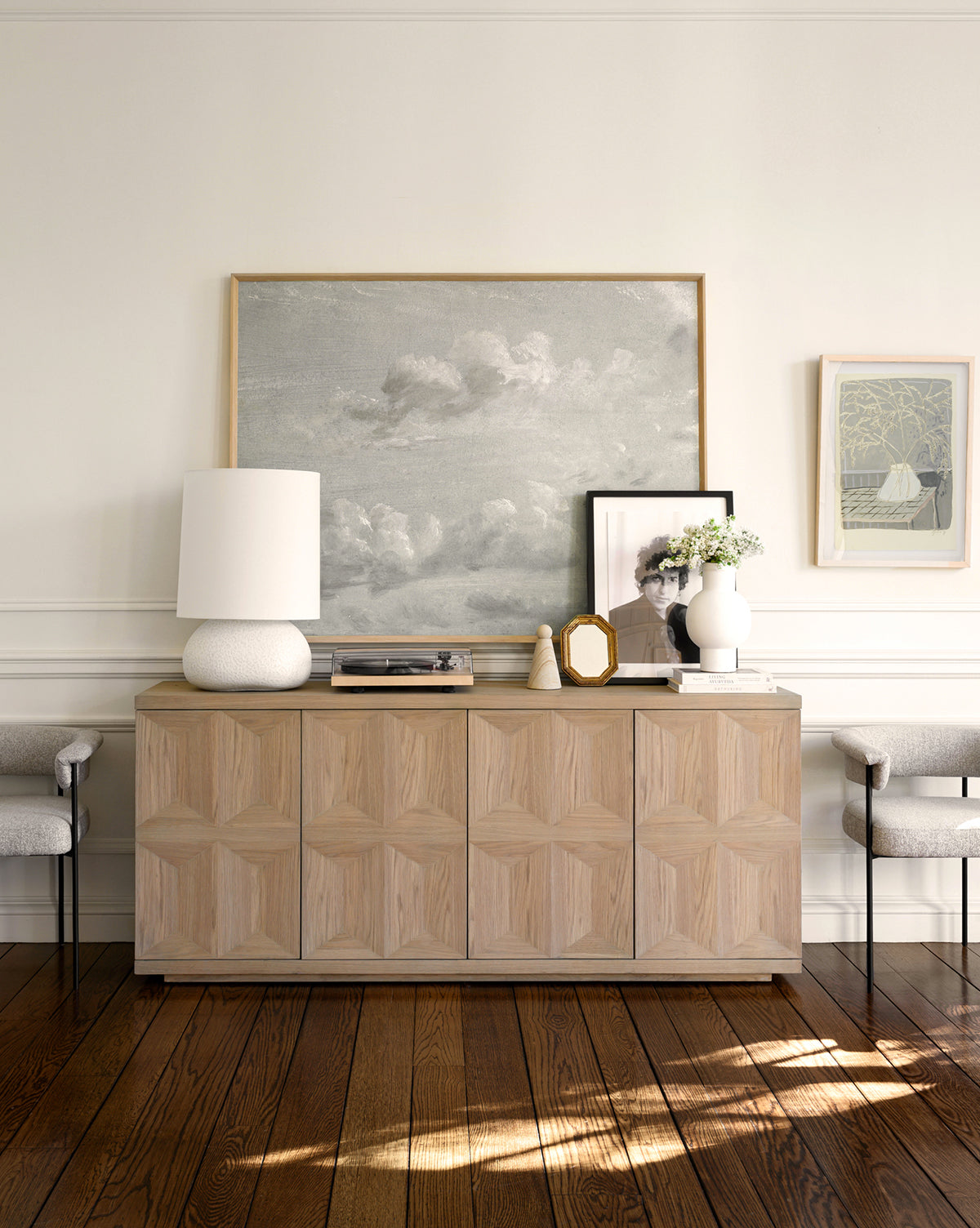A modern wood sideboard displays decor and a white lamp, while Gray Clouds wall art hangs above. Two upholstered chairs flank the setup, all resting on polished wood floors for an inviting, relaxed home atmosphere.