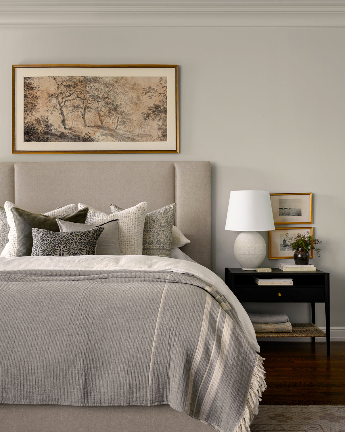 A cozy bedroom with the Mina Bed’s beige upholstered frame, layered gray and white pillows, a gray blanket, black nightstand with white lamp, art, fresh flowers, and a landscape painting above the bed, by McGee & Co.