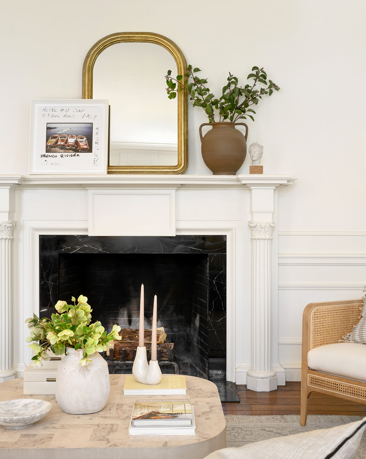 A cozy living room has a white fireplace with a gold-framed mirror, greenery in a soft brown vase, framed art, and a sculpture. The coffee table features candles and the Rounded Dual Handled Vase filled with flowers.