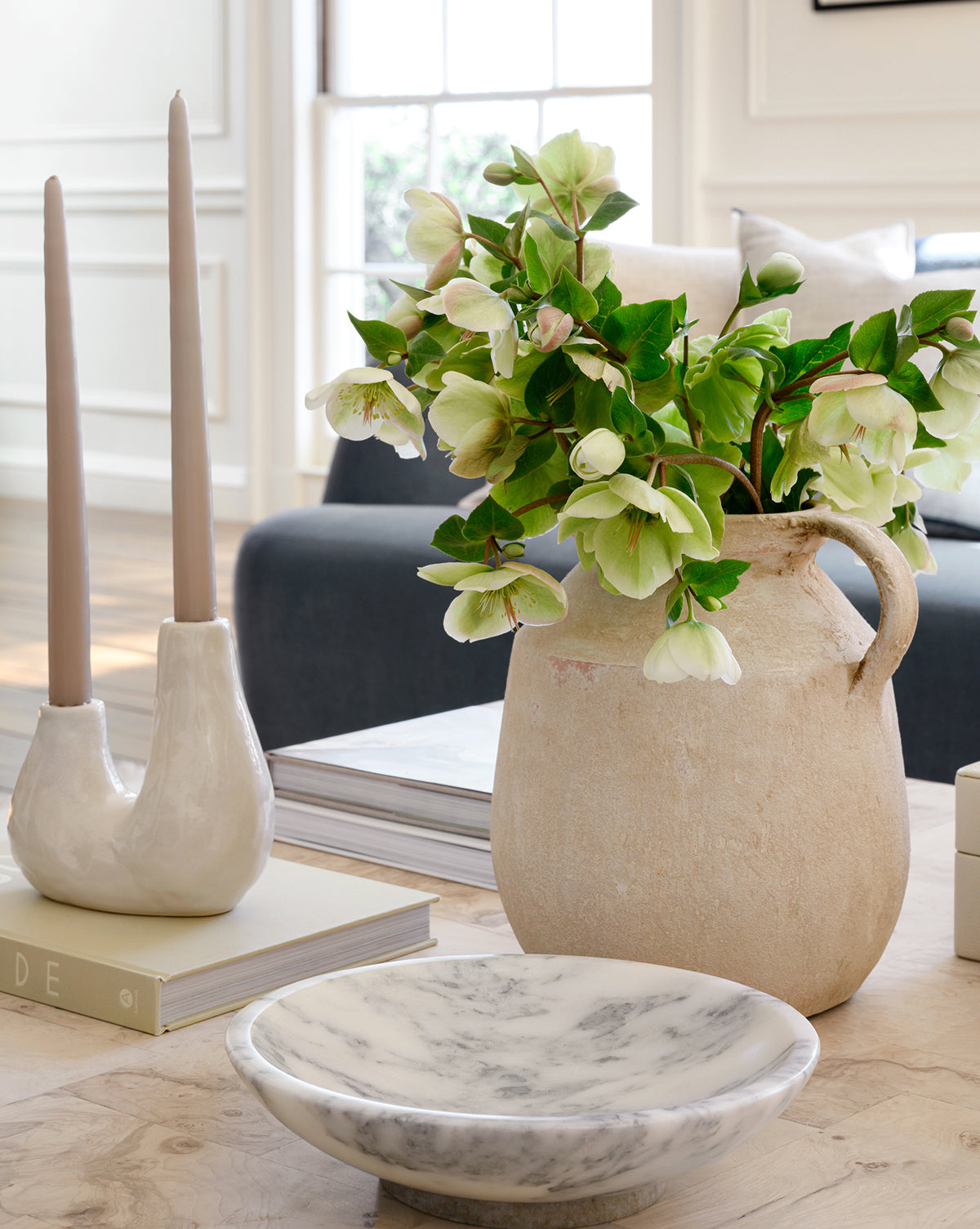 A rustic beige ceramic pitcher with green and white flowers sits on a wooden table beside a modern candle holder with two beige candles and the elegant Arabesco Marble Bowl, framed by sunlight from a window and a dark sofa in the background.