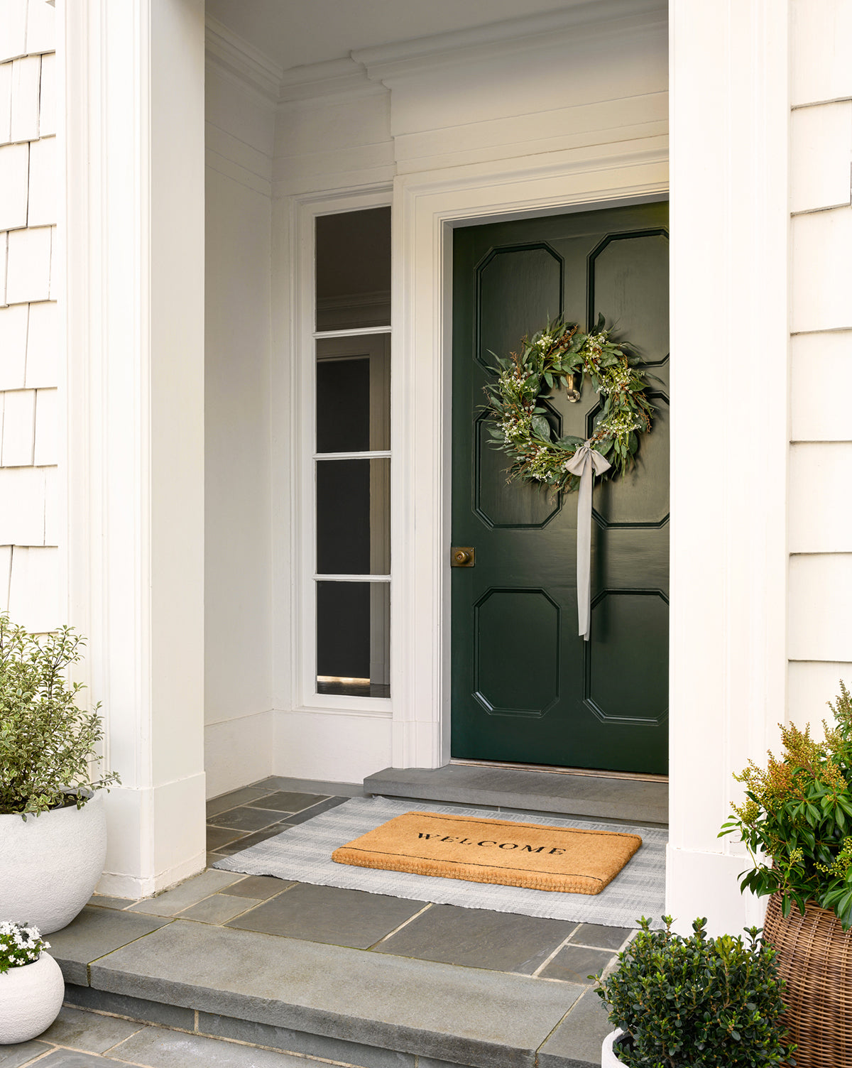 A green front door features a Faux Wild Grass & Eucalyptus Wreath with ribbon, flanked by tall windows. A “Welcome” doormat rests on the stone doorstep, surrounded by potted green plants on each side of the entryway.