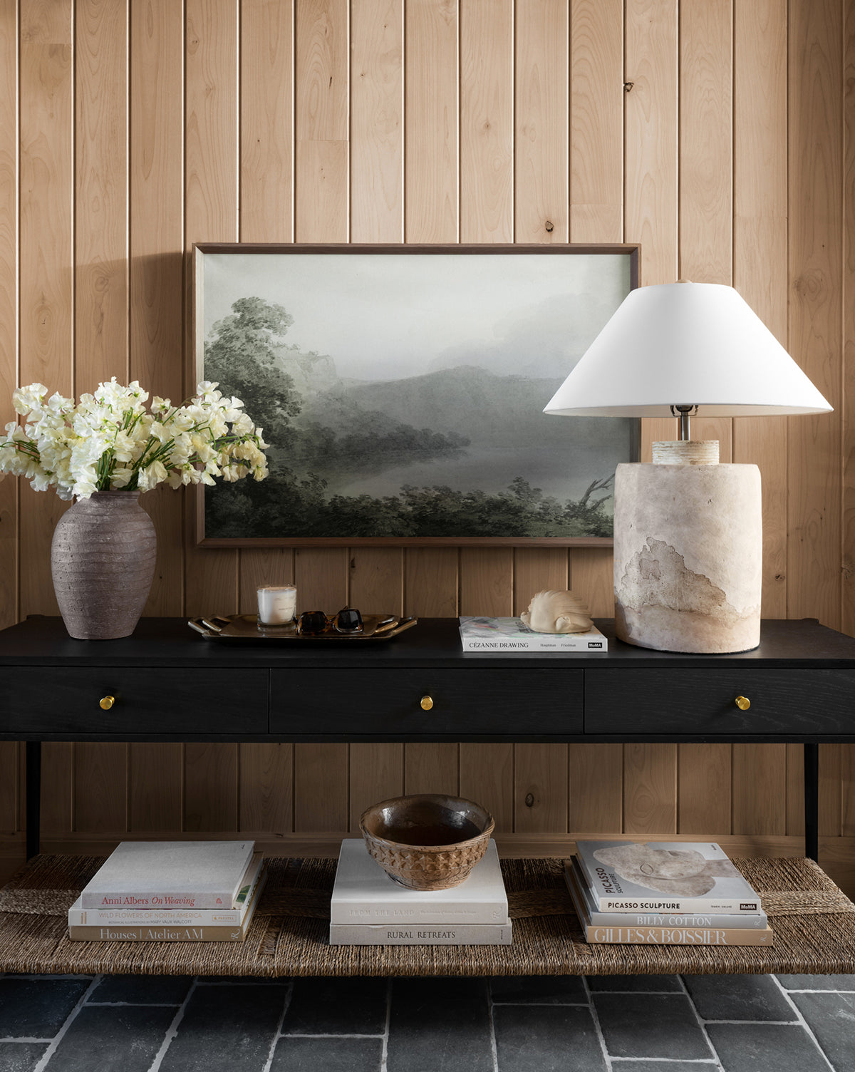 A black console table against a wood-paneled wall displays a vase of white flowers, the Verde Landscape giclee print, a stone lamp, books, and decor. Books and a bowl sit on the lower shelf over stone tile flooring.