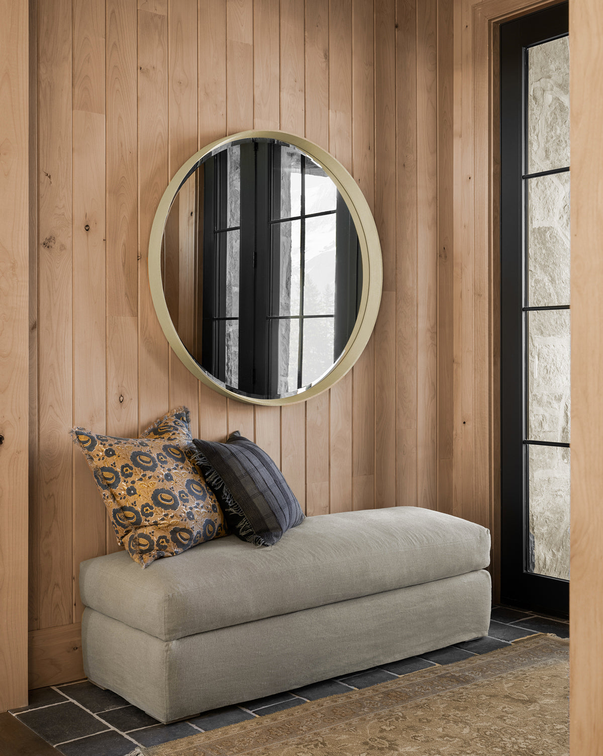 A cozy corner with light wood paneling, a round mirror, beige bench, and patterned pillows. Black-framed windows brighten the space, showcasing stylish living room furniture and the customizable Macy Slipcover Ottoman by Community—by McGee & Co.