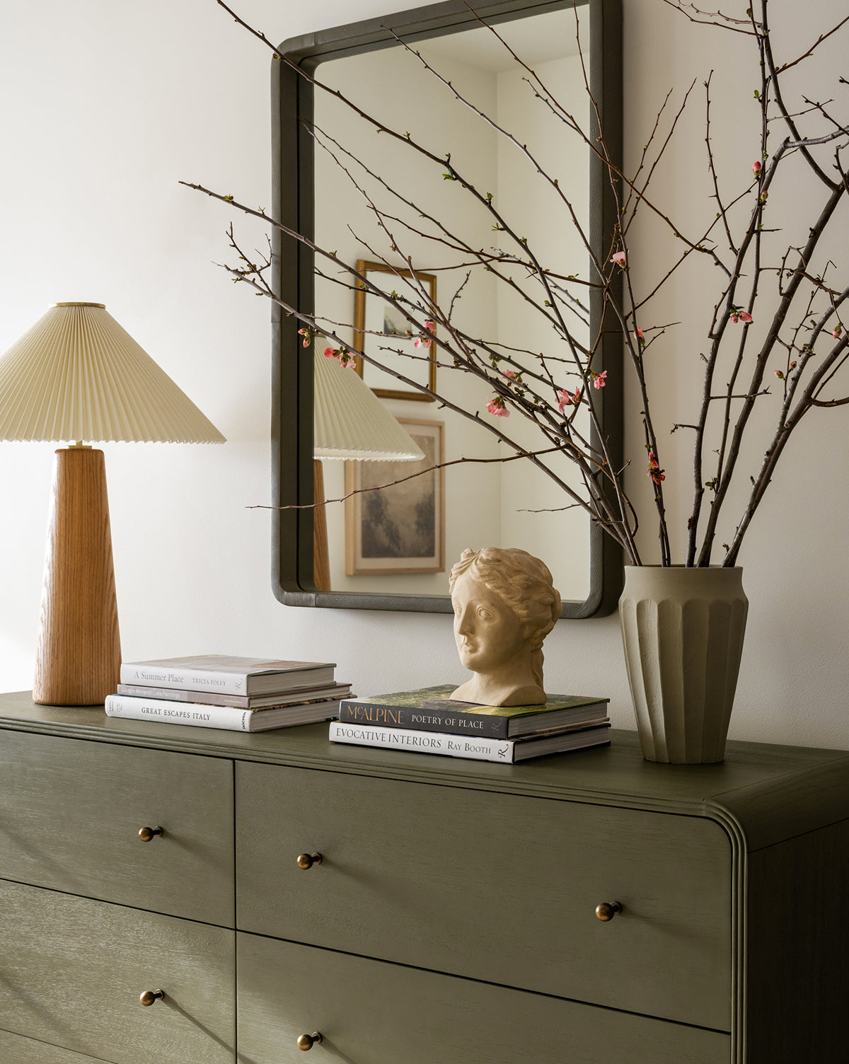 A green dresser with brass knobs holds a pleated lampshade, stacked books, the Hestia Bust sculpture, and a large vase of blossoming branches. A rectangular mirror is mounted above the dresser.