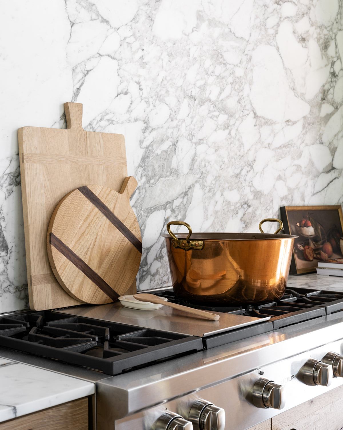 A copper pot sits on a stainless steel gas stove, with a Rectangle Bread Board in natural oak and walnut finish propped against a white marble backsplash. A small framed painting is in the corner, and a wooden spoon rests beside the pot, by McGee & Co.