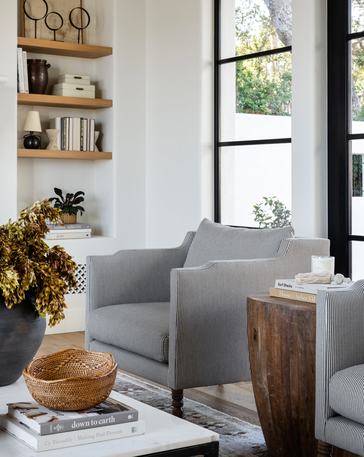 A cozy living room showcases the Gemma Lounge Chair (Ready to Ship) in premium fabric, paired with a round wooden side table, books, a woven basket, and a black vase with dried leaves. Shelves and large windows complete the inviting space by McGee & Co.