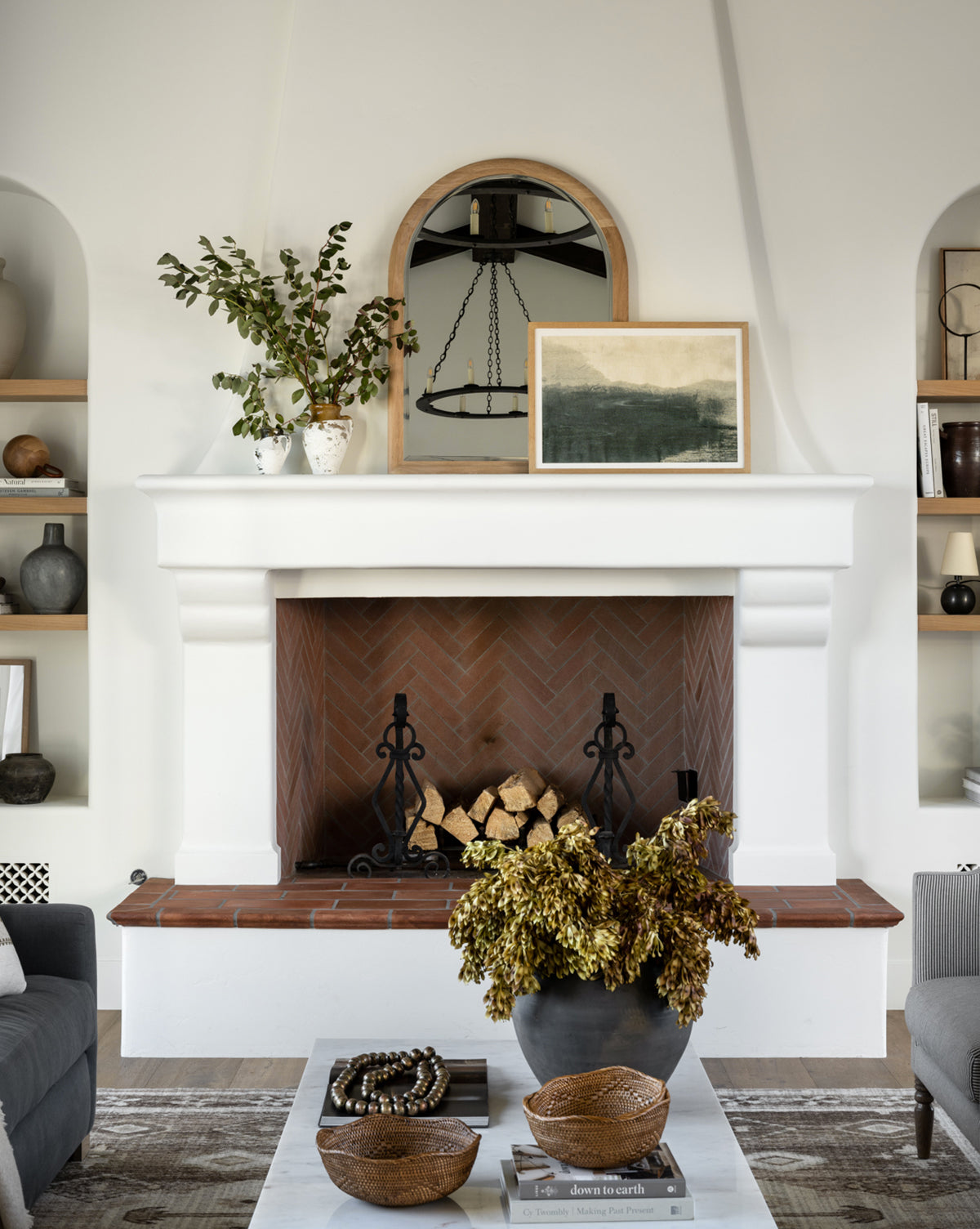 A stylish living room features a white fireplace with firewood, a Reta Arched Wall Mirror above the mantel, framed art and vases with greenery. Natural oak shelves hold books and decor; the coffee table displays bowls and a floral bouquet by McGee & Co.