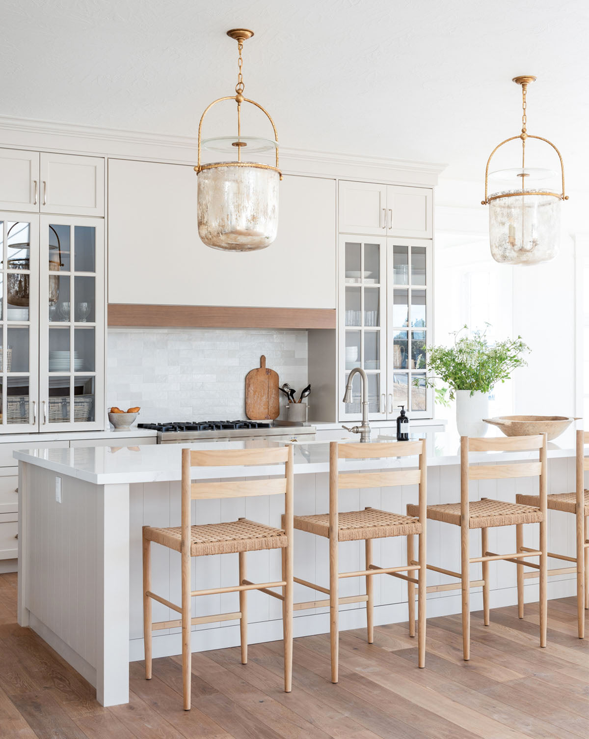 A bright, modern kitchen with white cabinets, a large island with a white countertop, three Eloise Woven Counter Stools, glass pendant lights, and a vase with greenery. Light wood floors complete the airy look, by McGee & Co.