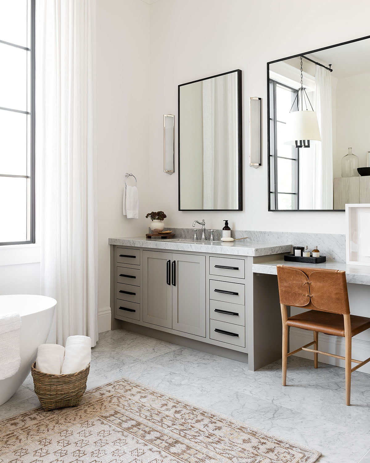 A modern bathroom features a freestanding bathtub, light gray double-sink vanity, large mirrors, brown leather chair, marble floors, and the Anya Neutral Hand-Knotted Wool Rug paired with a basket of rolled towels for added comfort, by McGee & Co.
