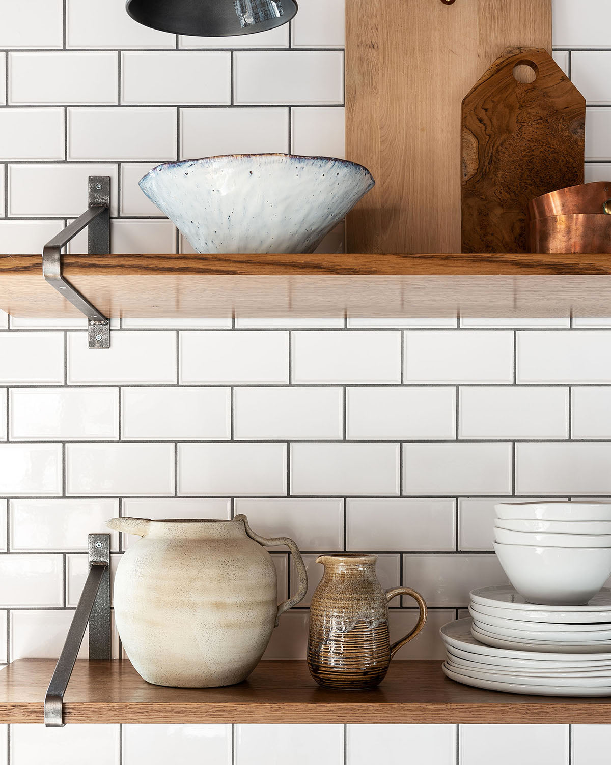 Open wooden shelves display ceramic bowls, plates, a Weathered Ceramic Jug, and pitchers against a white subway tile backsplash. A cutting board and copper pot on the upper shelf enhance the kitchen’s vintage style.