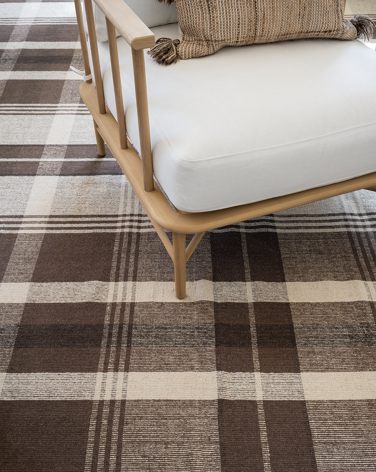 A light wooden armchair with white cushions sits on the Orton Handwoven Wool Rug, which features a brown and beige plaid pattern. A textured striped pillow with tassels is placed on the chair, by McGee & Co.