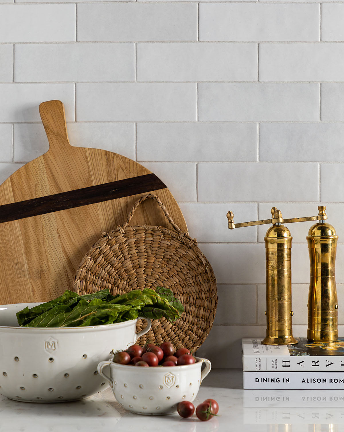 A kitchen counter features the Round Oak Bread Board, a woven trivet, two brass pepper mills, ceramic bowls with leafy greens and cherry tomatoes, and stacked cookbooks against a white tiled backsplash, by McGee & Co.