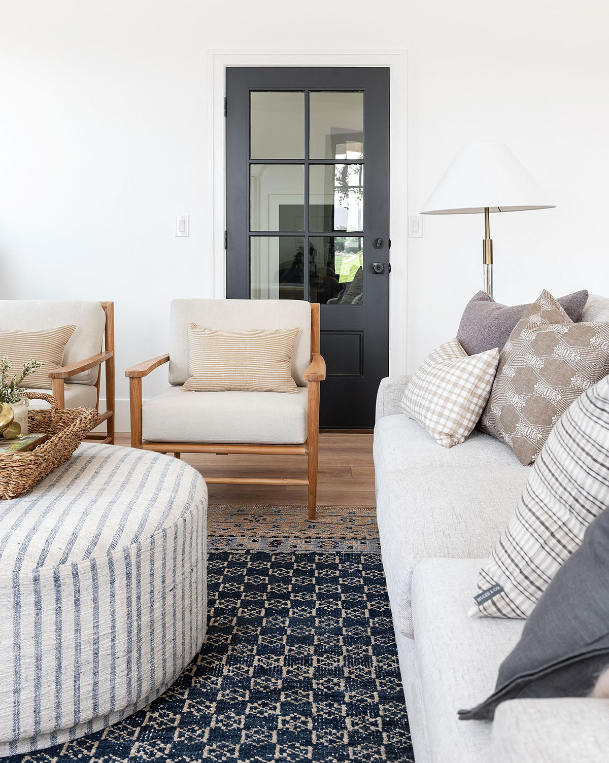 A bright, modern living room featuring a black door, beige armchair, light gray sofa with patterned pillows, striped ottoman, and tall floor lamp is anchored by the Anya Indigo Hand-Knotted Wool Rug for added elegance, by McGee & Co.