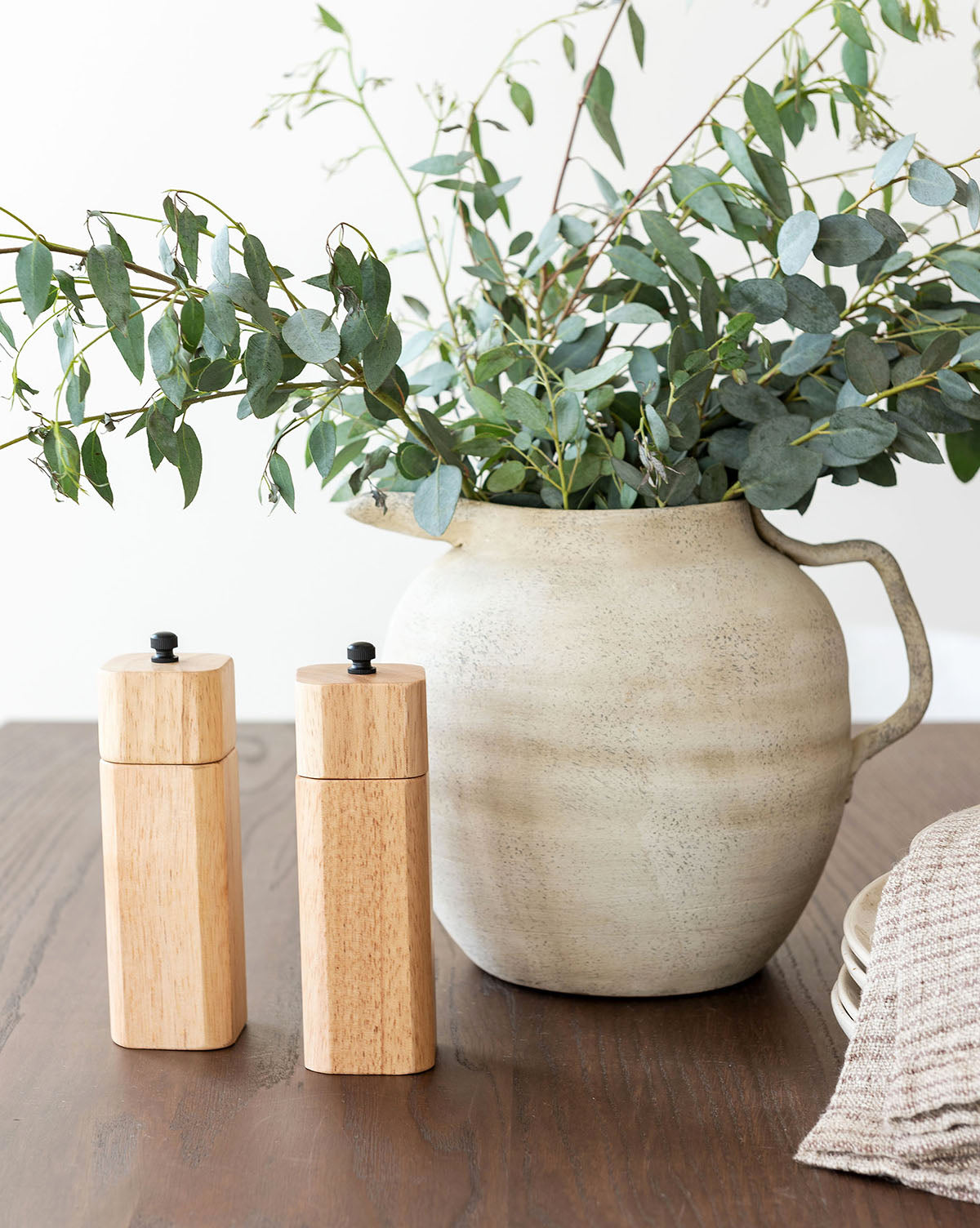 A Weathered Ceramic Jug filled with leafy green branches sits on a wooden table beside two wooden salt and pepper mills, a folded beige cloth, and stacked white plates.