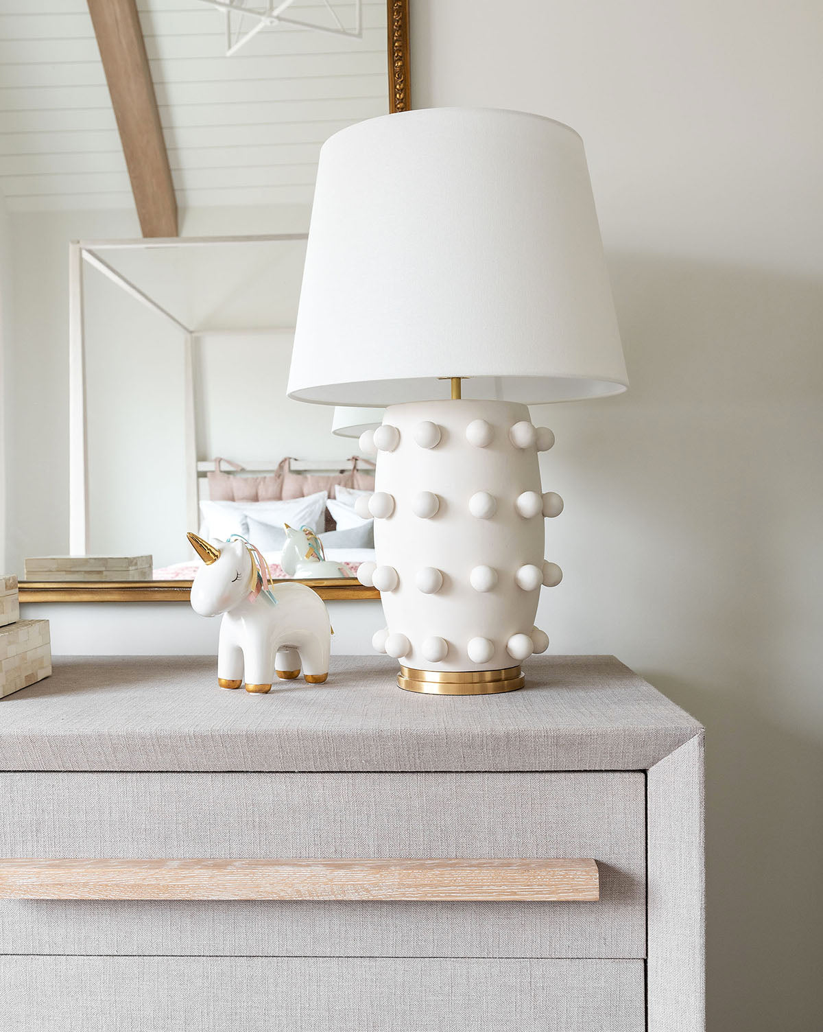 A dresser holds the Linden Table Lamp with a linen shade and gold accents, alongside a small ceramic unicorn figurine; in the background, part of a bed and a mirror are visible, by McGee & Co.