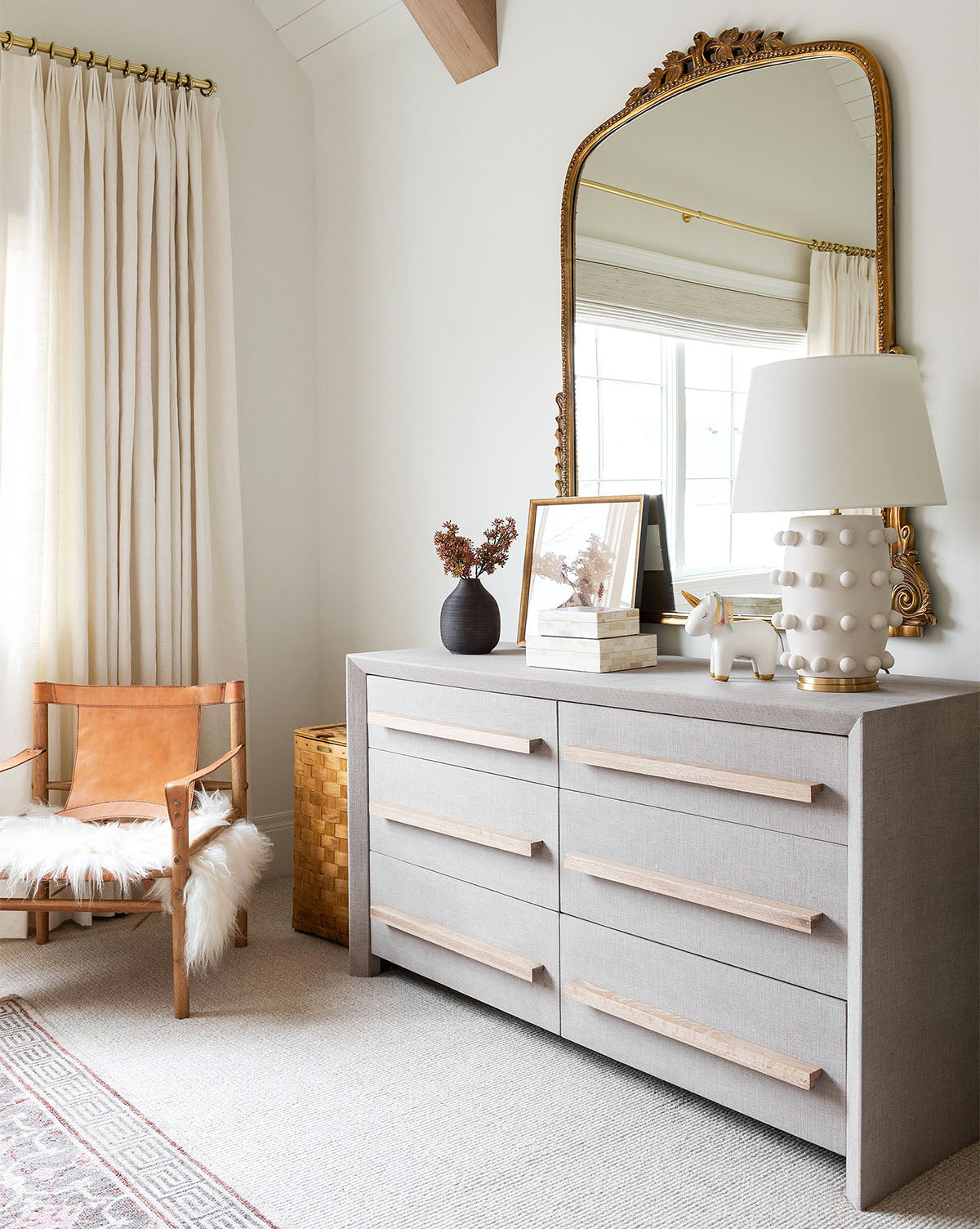 A modern bedroom corner features the Linden Table Lamp with a linen shade on a gray dresser, paired with a large ornate gold mirror, tan leather chair with white throw, and light curtains softly covering the window by McGee & Co.
