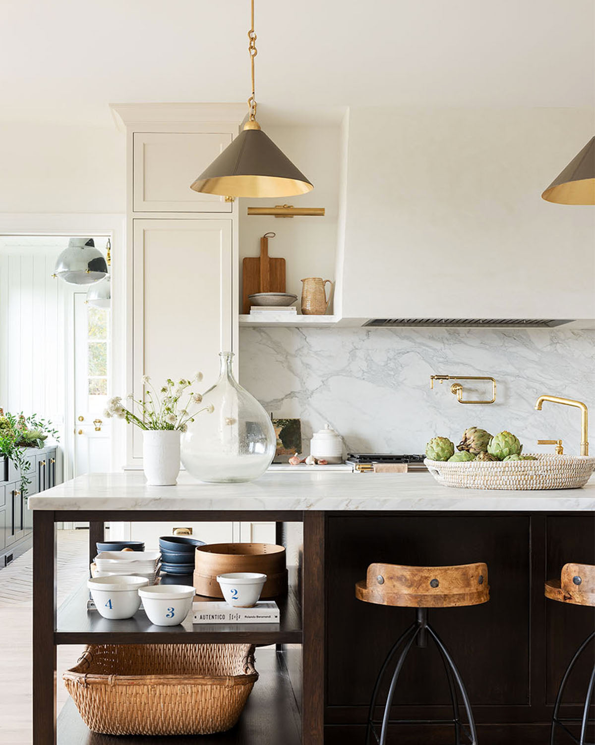 A bright, modern kitchen with a marble backsplash, gold fixtures, a dark wood island with open shelves, two wooden stools, and elegant Scarlett Pendants illuminating the counter by McGee & Co.