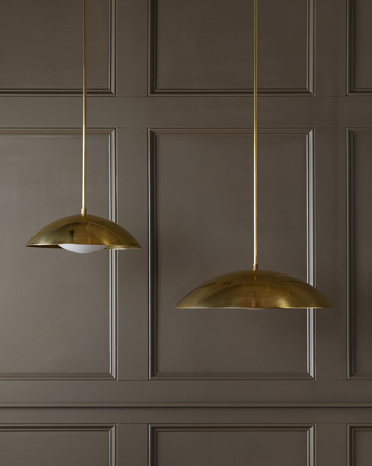 Two modern brass Aldorno Pendants hang before a brown paneled wall: the left exposes its bulb, while the right showcases a wide, round shade that fully conceals the bulb, by McGee & Co.